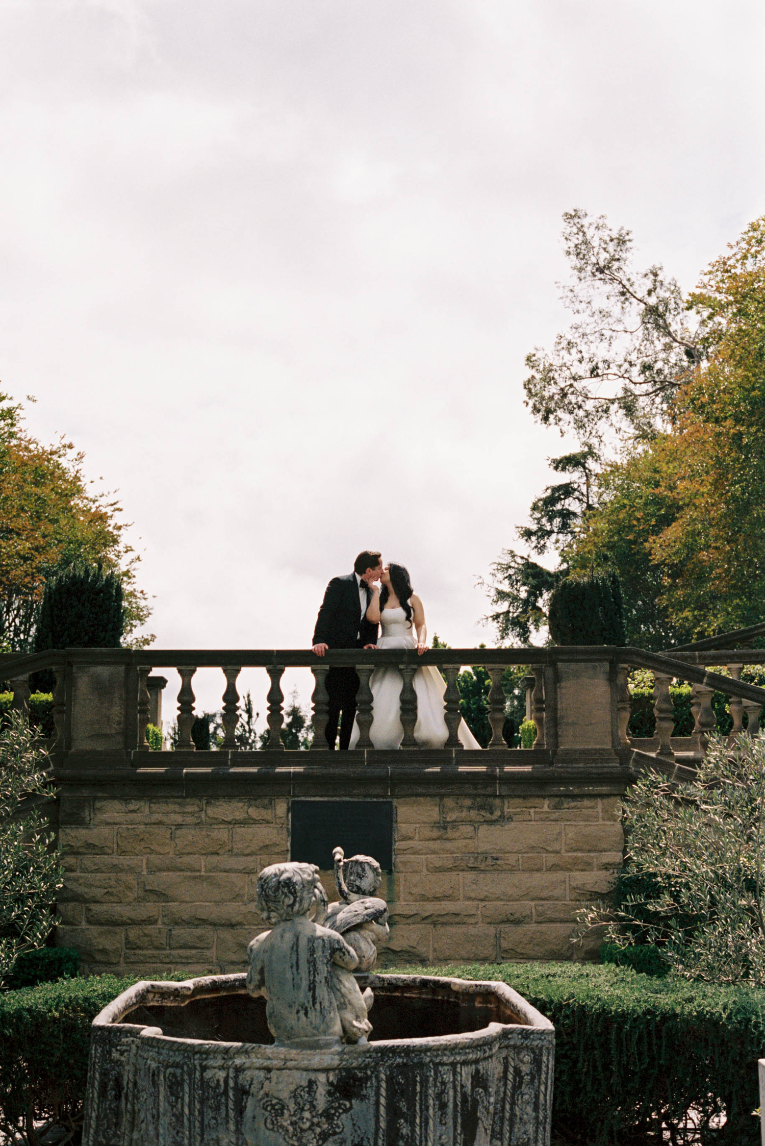 A wedding couple shares a kiss on a stone balustrade above a cherub fountain in an elegant garden setting.