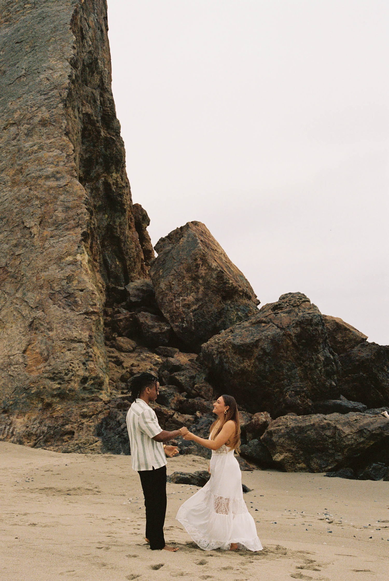 A couple holds hands and dances on a sandy beach with dramatic rocky cliffs in the background.