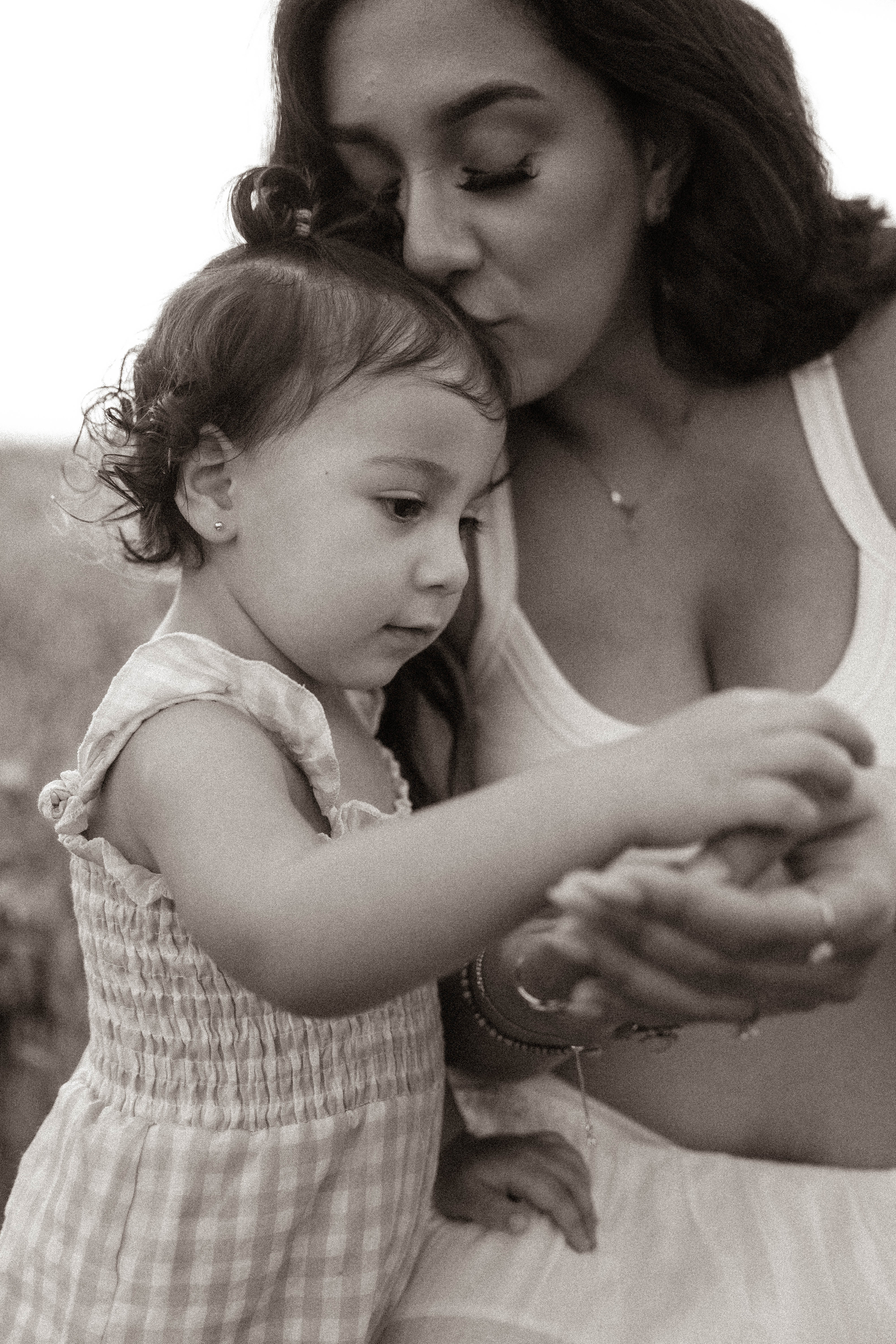 Black and white photo of a mother kissing her toddler daughter's head while they examine something in her hands.