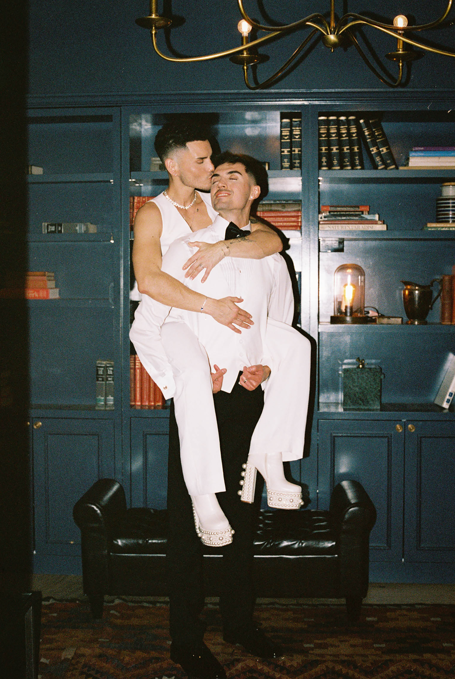 Two people in white outfits pose together in a dark blue library setting with brass chandelier.