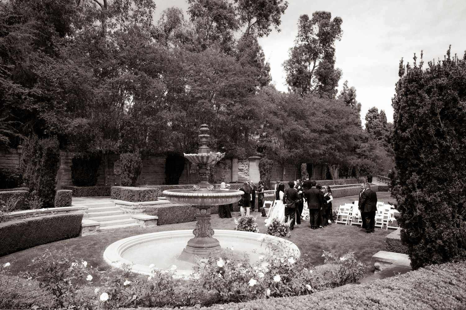 Black and white photo of an elegant garden wedding venue with a stone fountain centerpiece surrounded by lush trees.