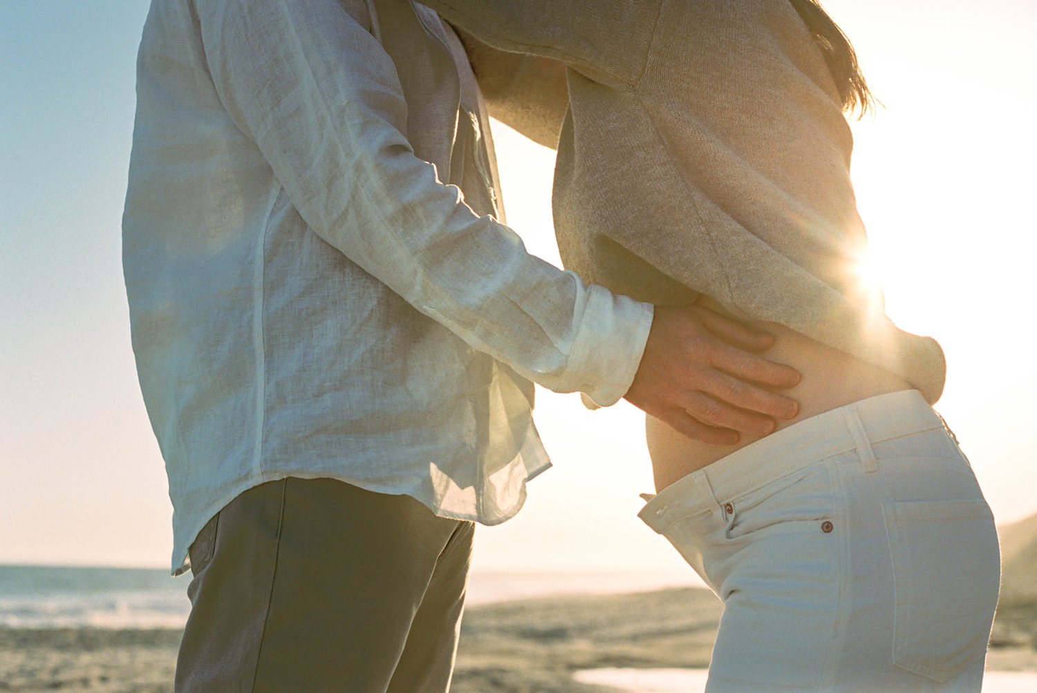 Couple embracing on a sunny beach at sunset, man touching pregnant woman's baby bump by the ocean.