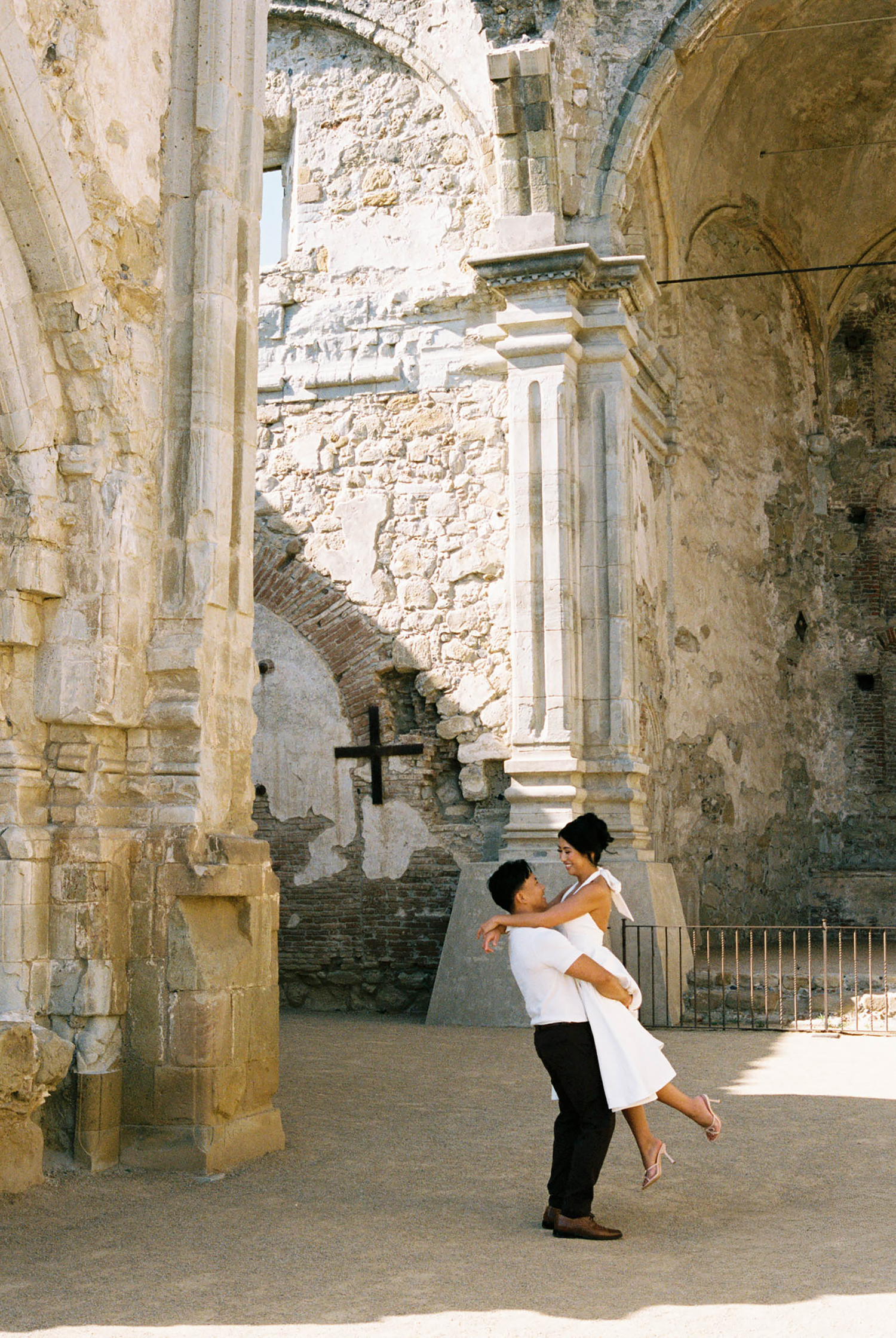 Couple embracing in romantic dance among ancient stone ruins with arched doorways and a cross.