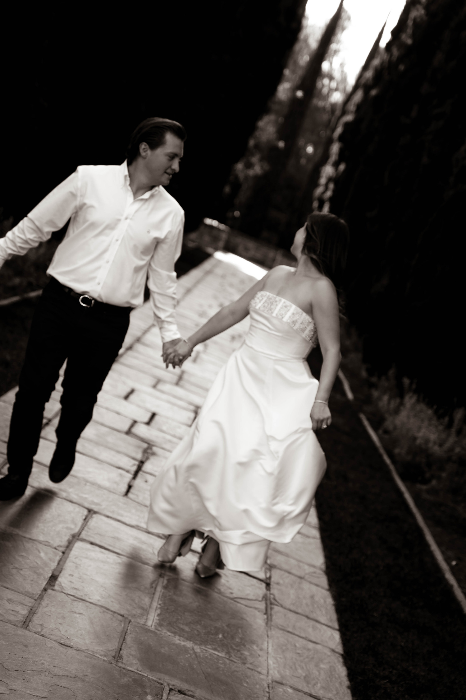 Black and white image of couple holding hands and walking along stone path lined with tall trees. Greystone Mansion & Gardens