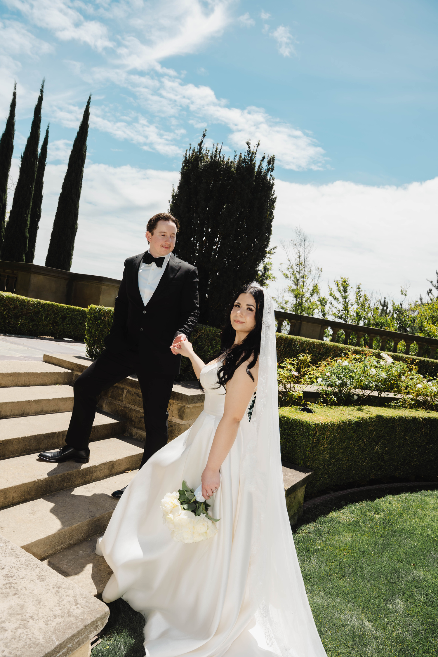 Bride in white gown holding bouquet leads groom in black tuxedo down stone steps in lush garden setting.