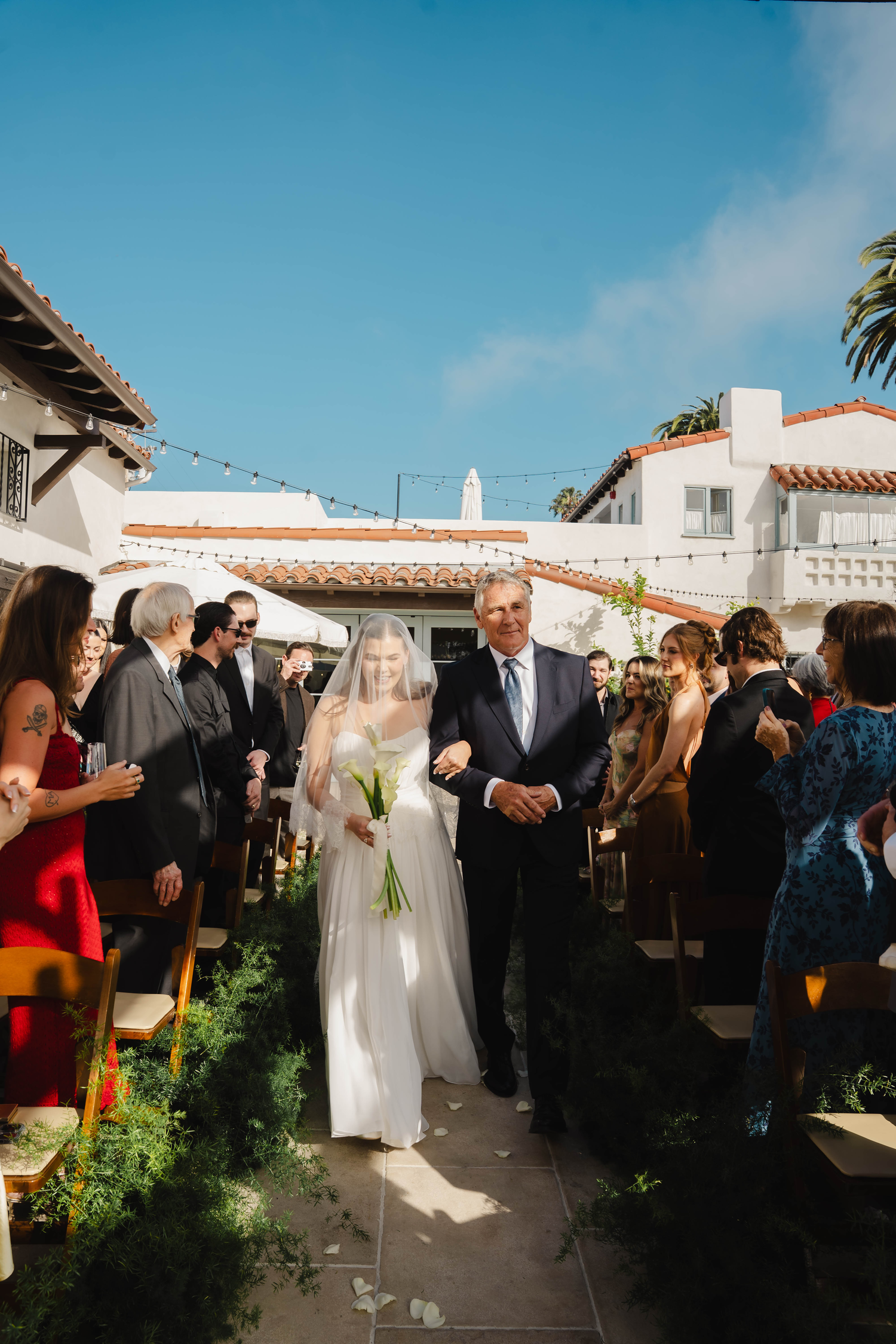 Bride walks down outdoor aisle with father on sunny day at elegant Spanish-style wedding venue.