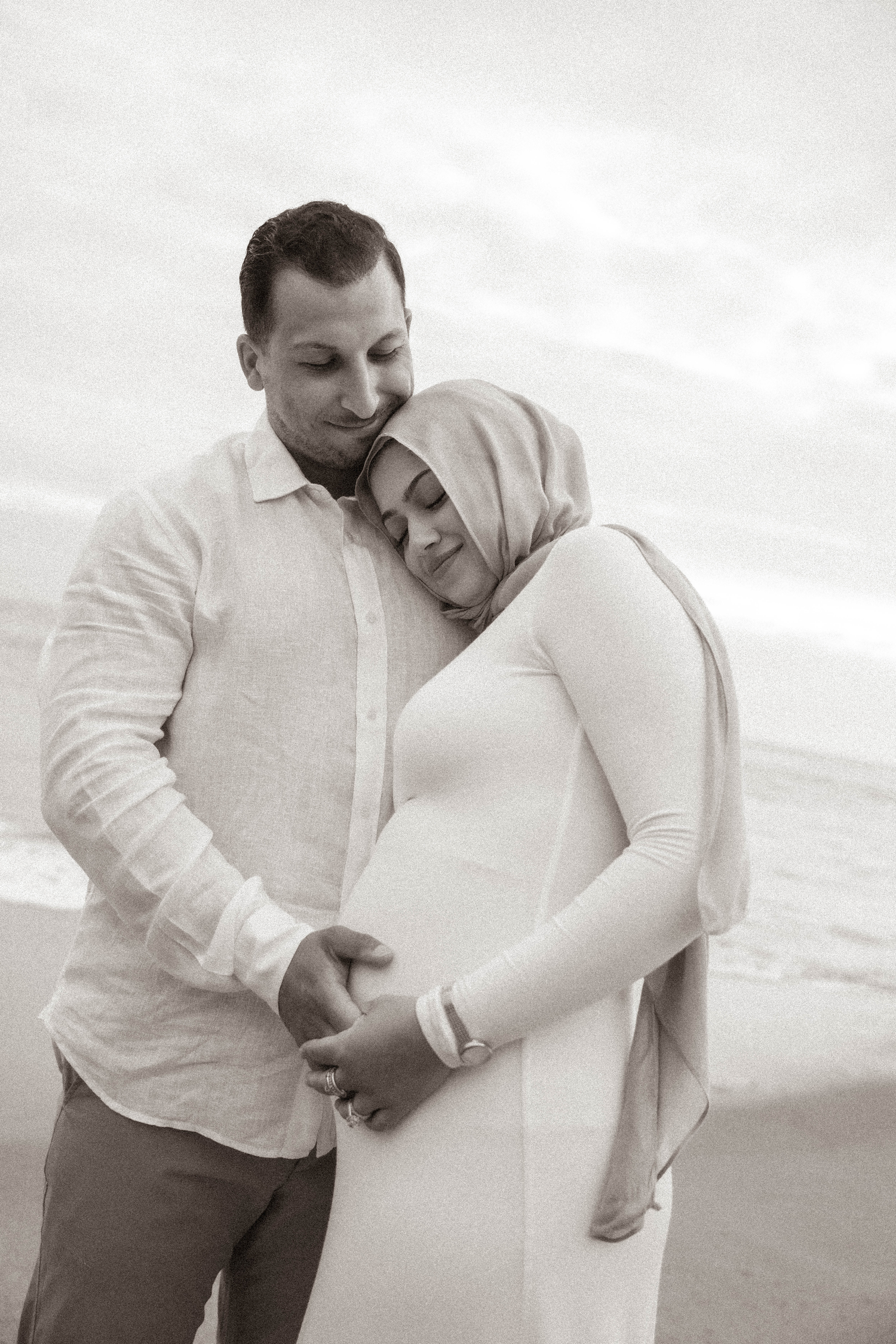 Black and white photo of a pregnant couple embracing on a beach, woman in hijab leaning on mans shoulder.