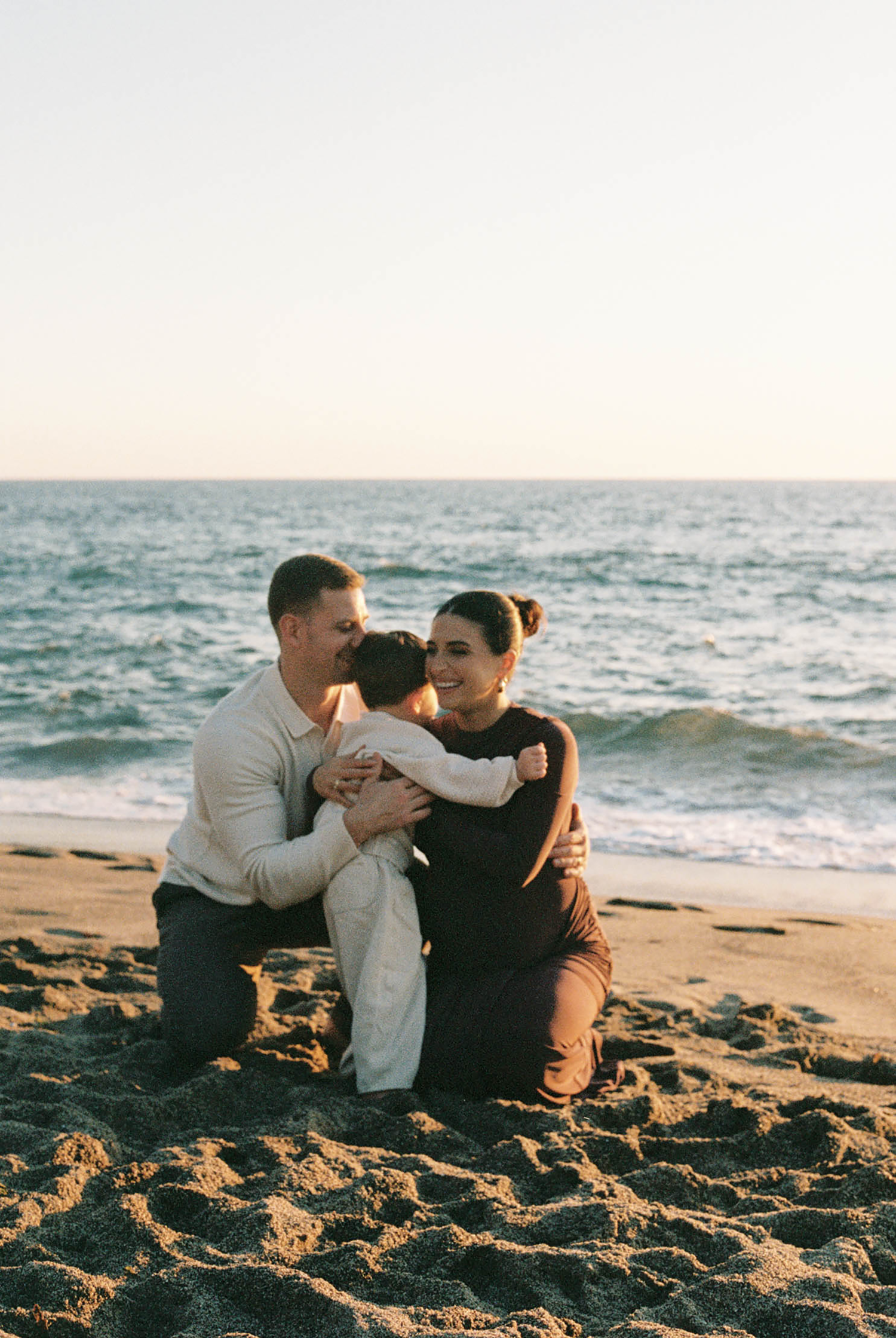 A couple kneels on a sandy beach at sunset, lovingly embracing their young child with ocean waves in the background.