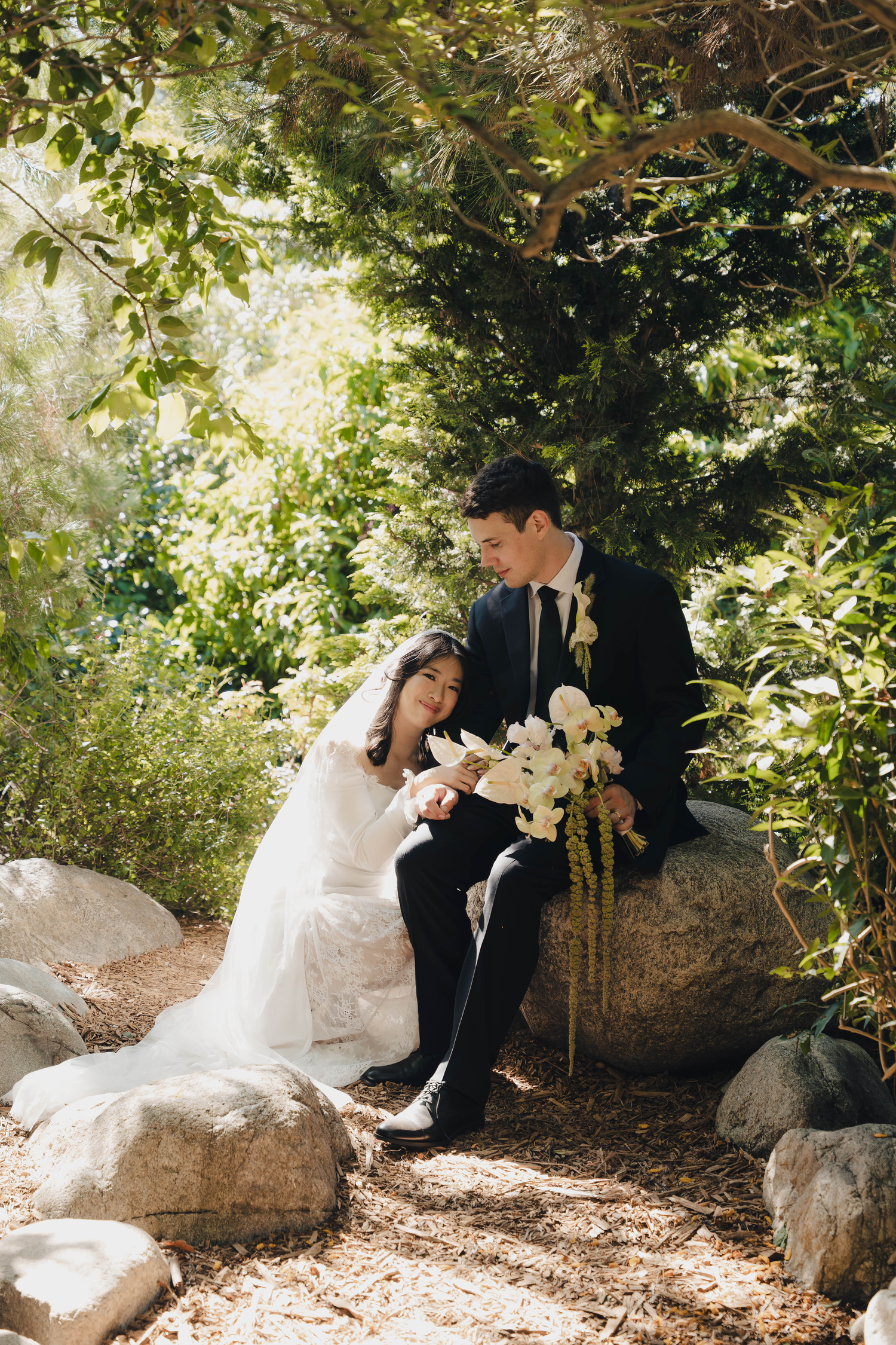 A bride and groom sit together on rocks in a lush garden, sharing a tender moment with white floral bouquet.