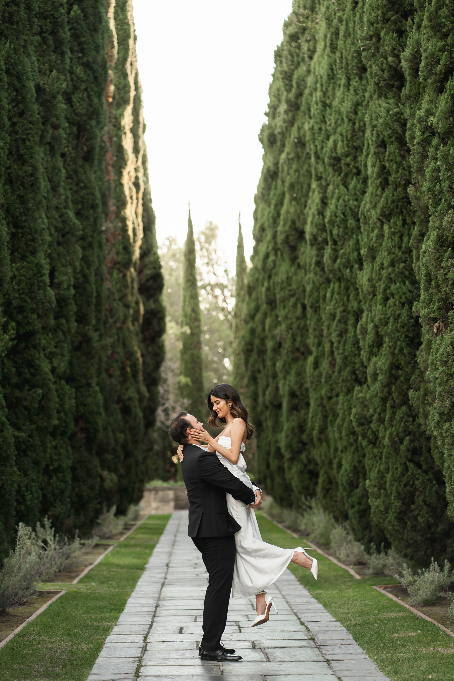 Groom lifts bride in white dress along cypress tree-lined garden path at outdoor wedding.