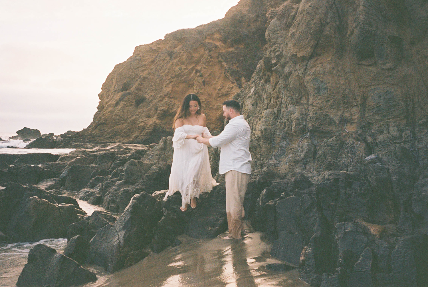 Couple holding hands on rocky beach during golden hour maternity photo session.