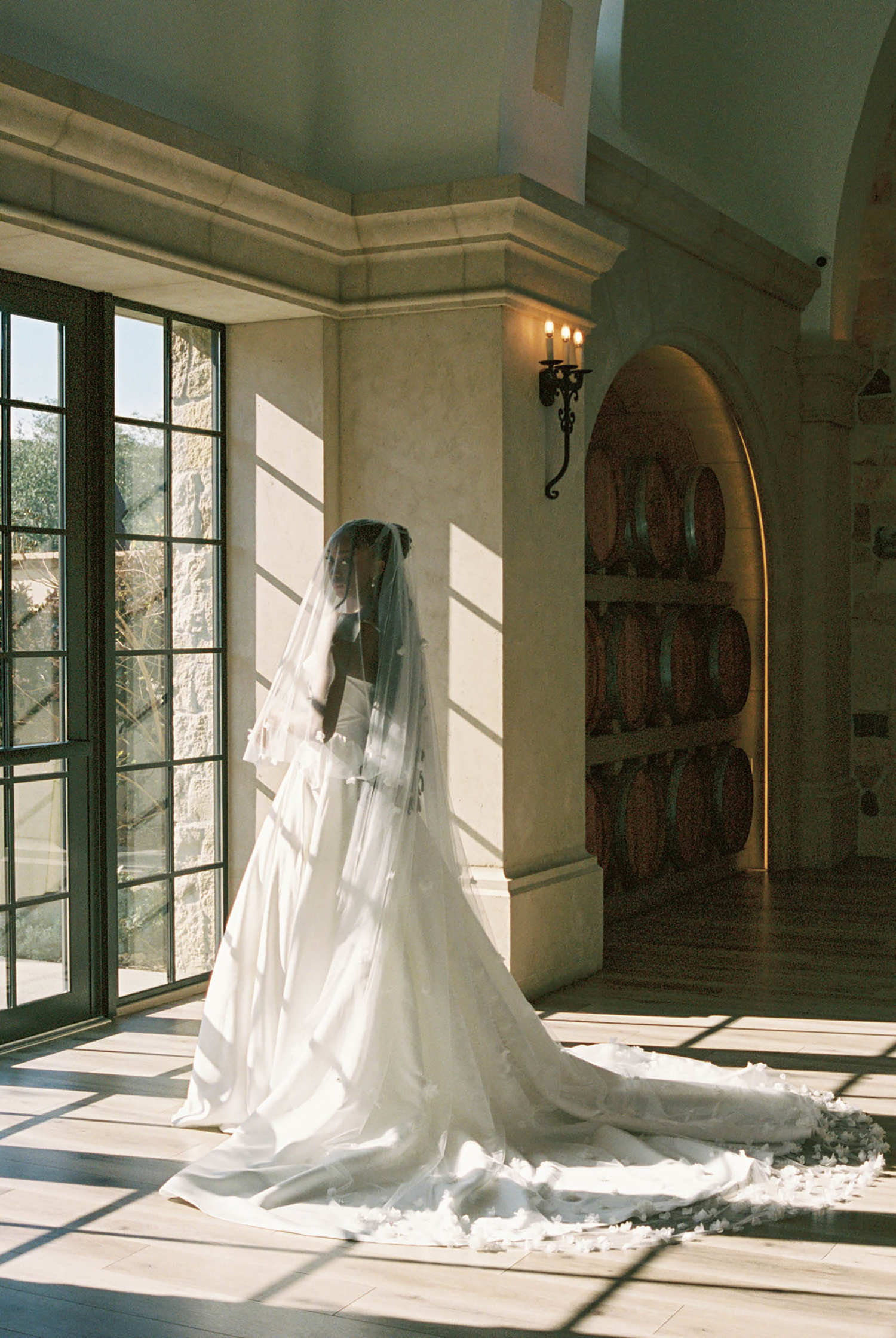 Bride in elegant white wedding gown and veil stands by tall windows in a sunlit venue with arched wine barrel alcove.