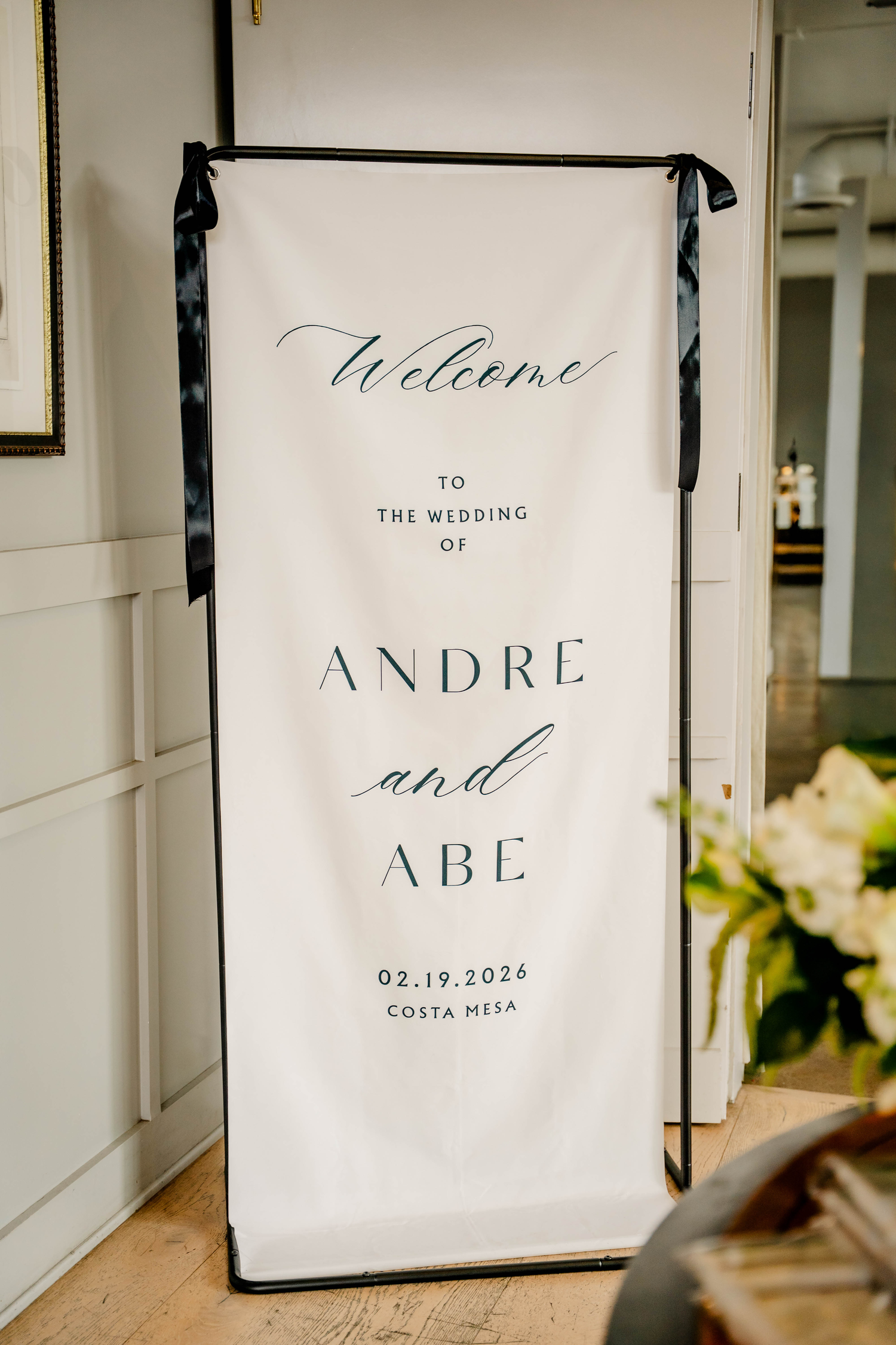 Elegant wedding welcome sign for Andre and Abe, dated 02.19.2026 in Costa Mesa, displayed on a black stand.