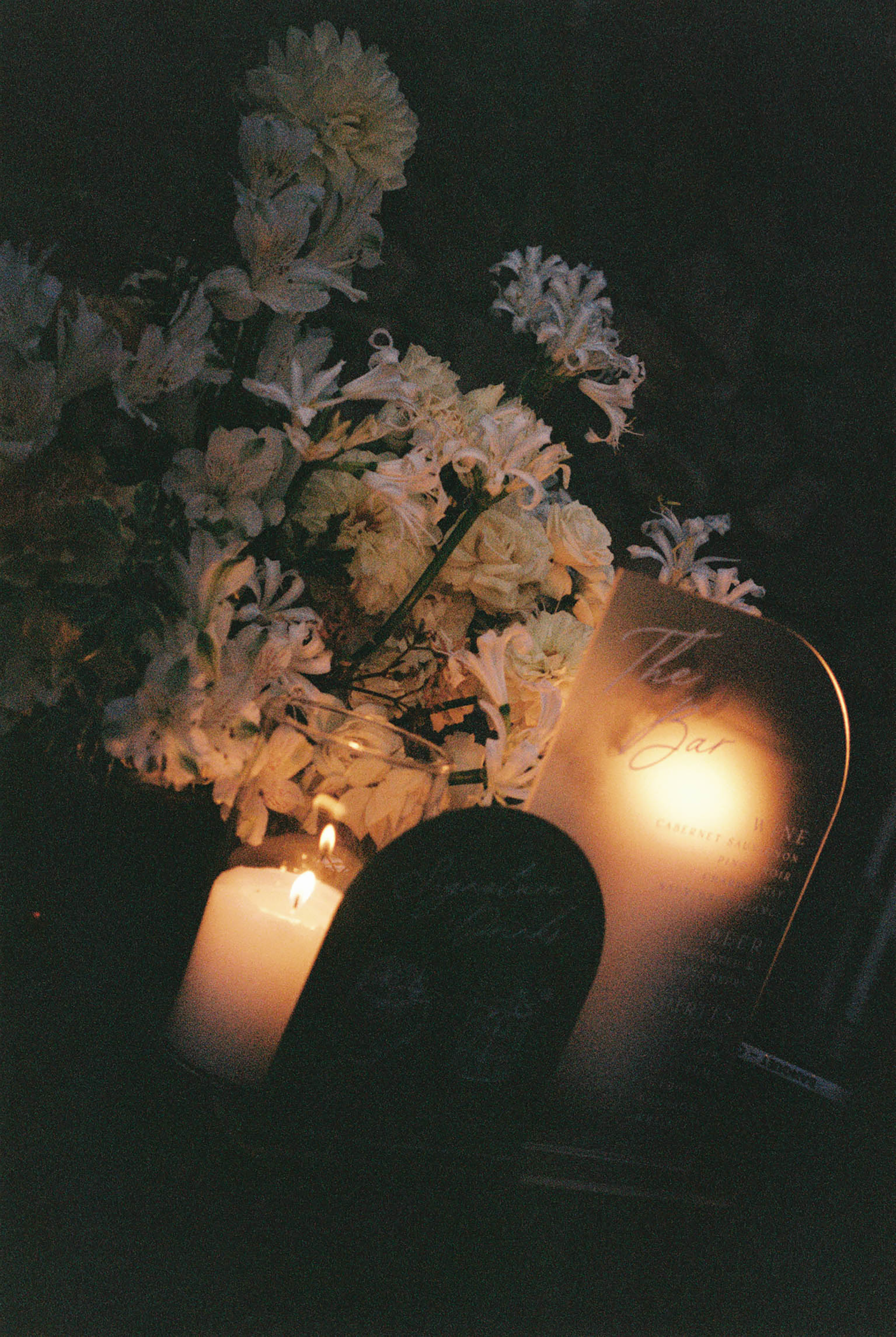 Moody candlelit wedding table display with white flowers, glowing candles, and decorative signage in dark setting.