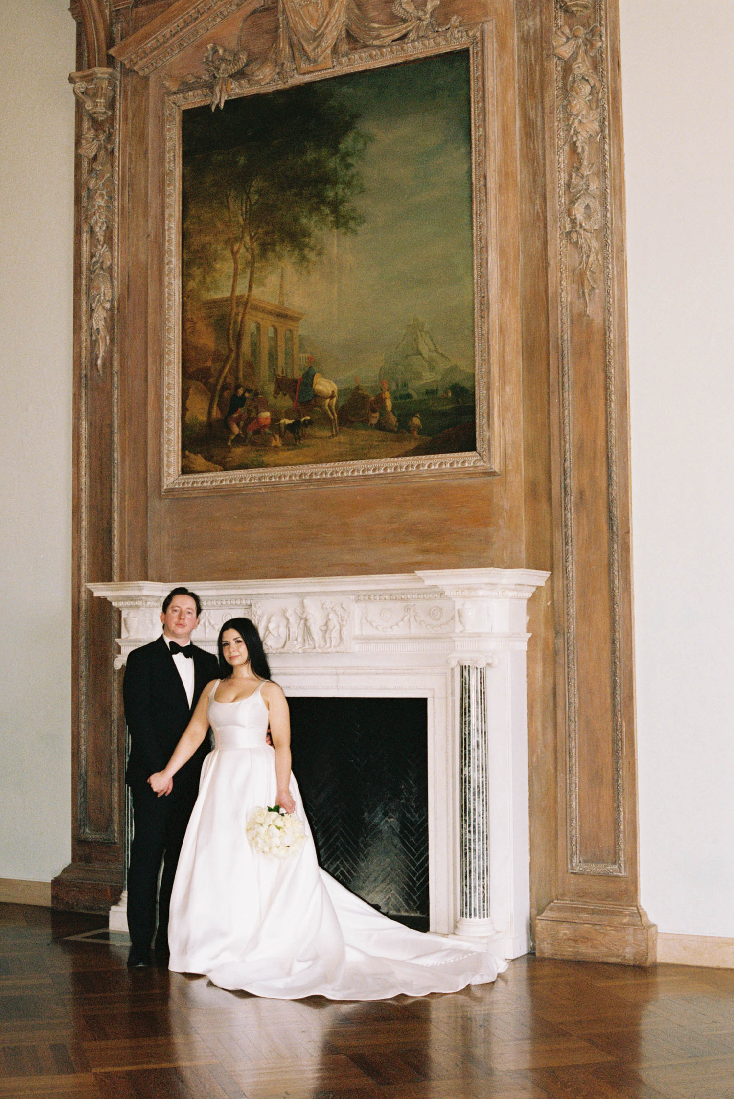 Bride and groom posing elegantly by a white fireplace beneath a large classical painting in a grand historic venue.