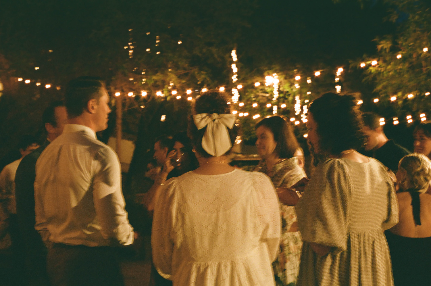 Outdoor evening wedding reception with string lights, guests in white dresses dancing under glowing bulbs in a garden setting.