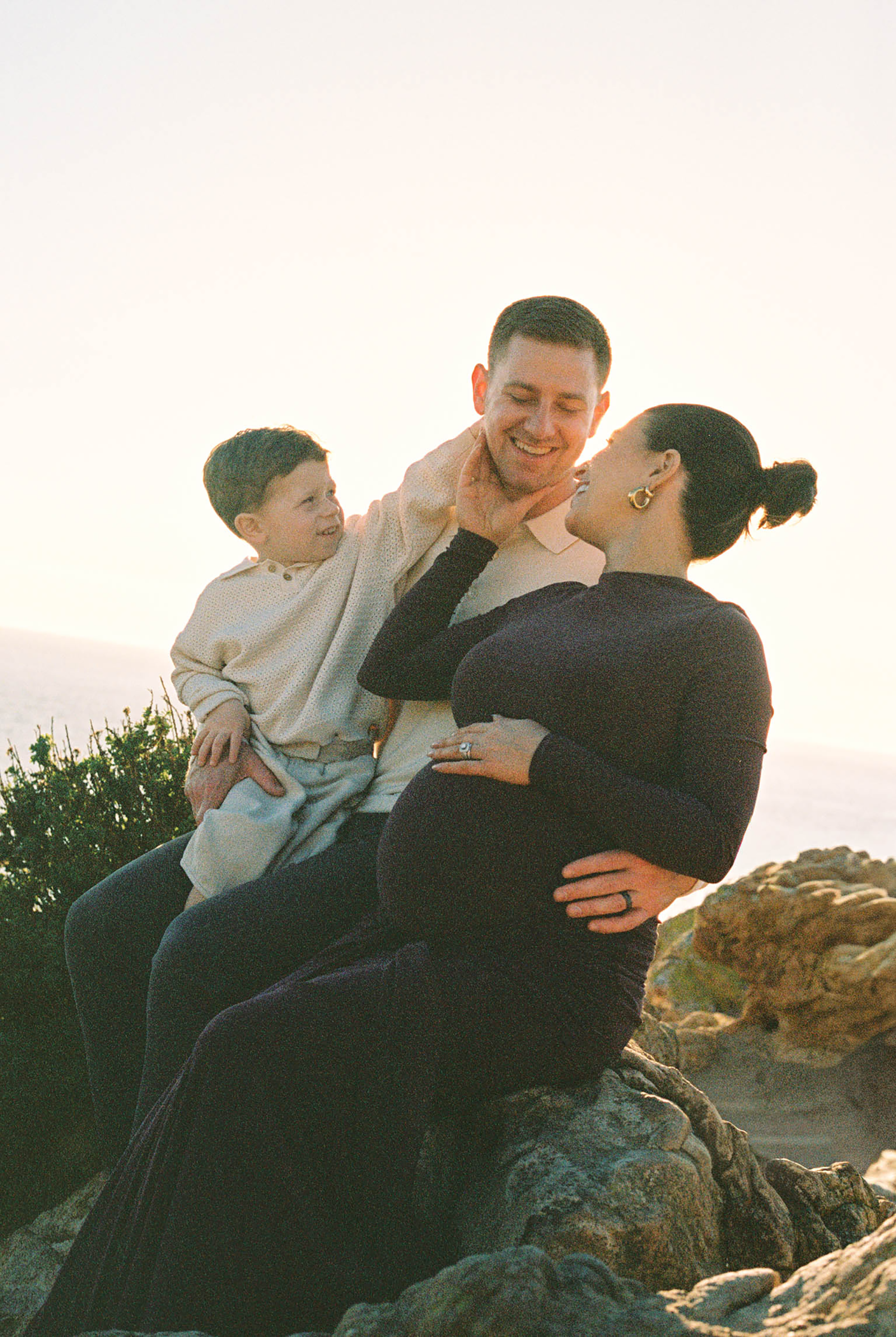 A pregnant woman sits on rocks with her partner and young son during a golden hour outdoor maternity photo session.