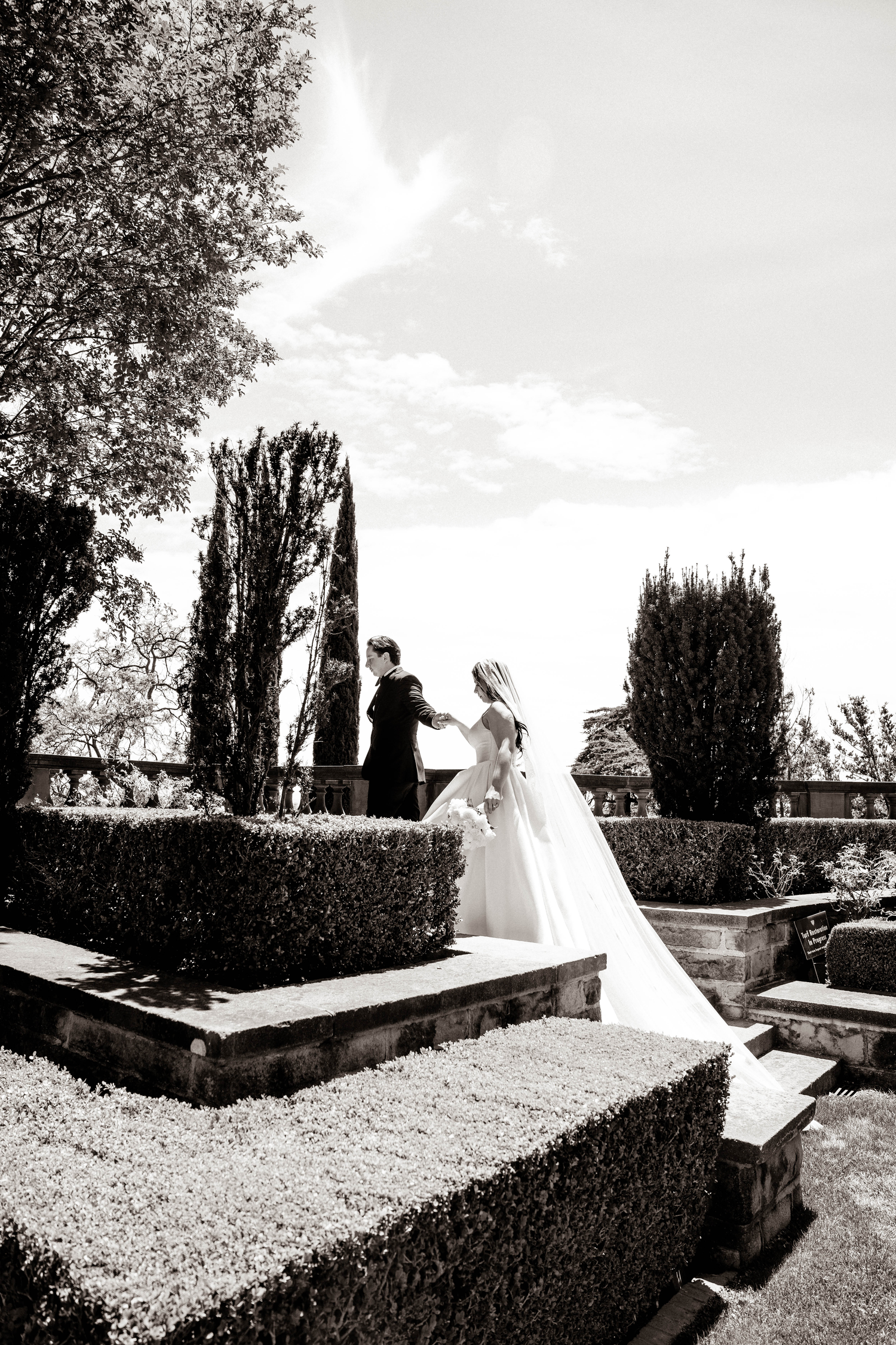Black and white wedding photo of bride and groom on garden steps surrounded by manicured hedges and cypress trees.