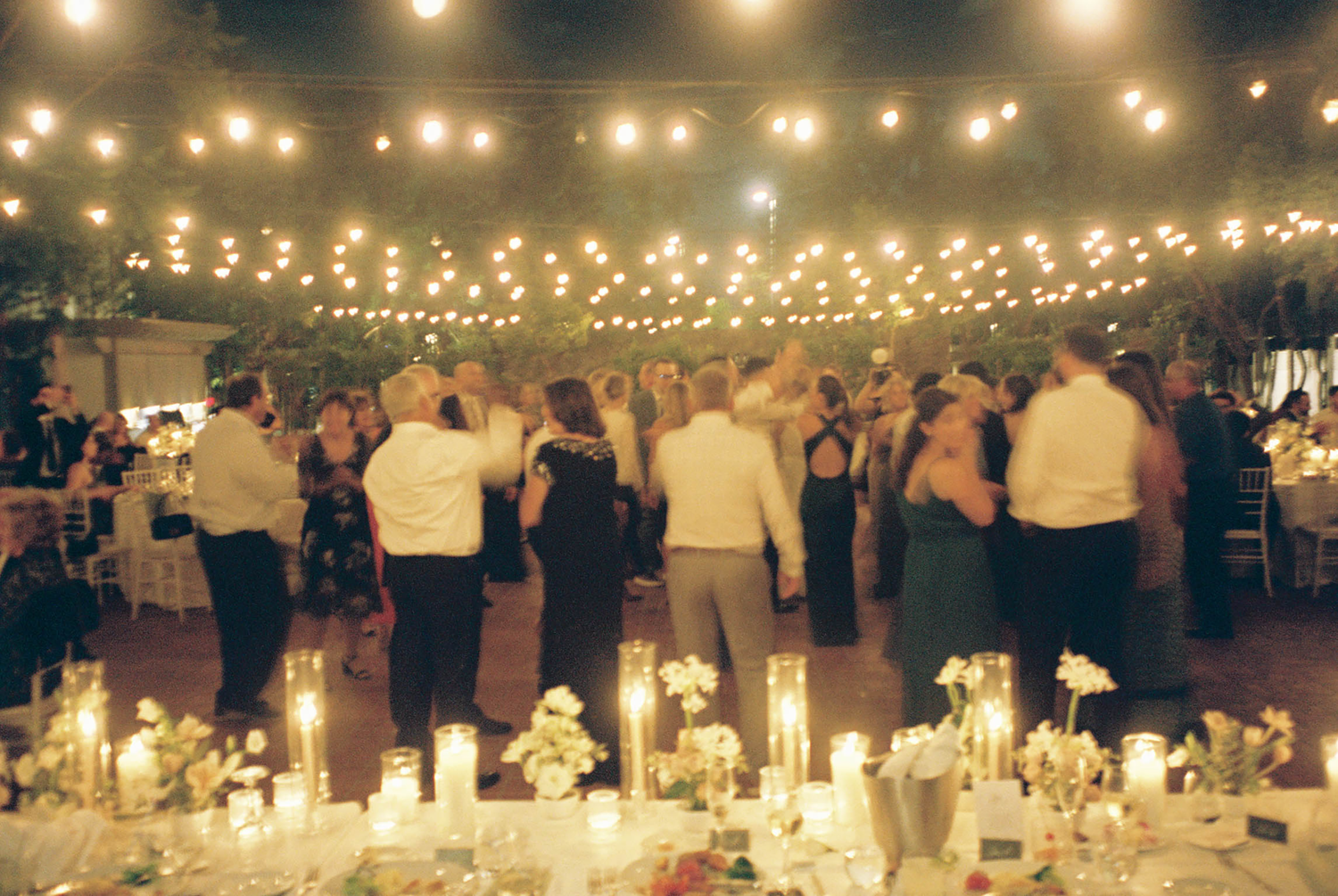 Guests dancing under warm string lights at an elegant outdoor wedding reception with candlelit tables.