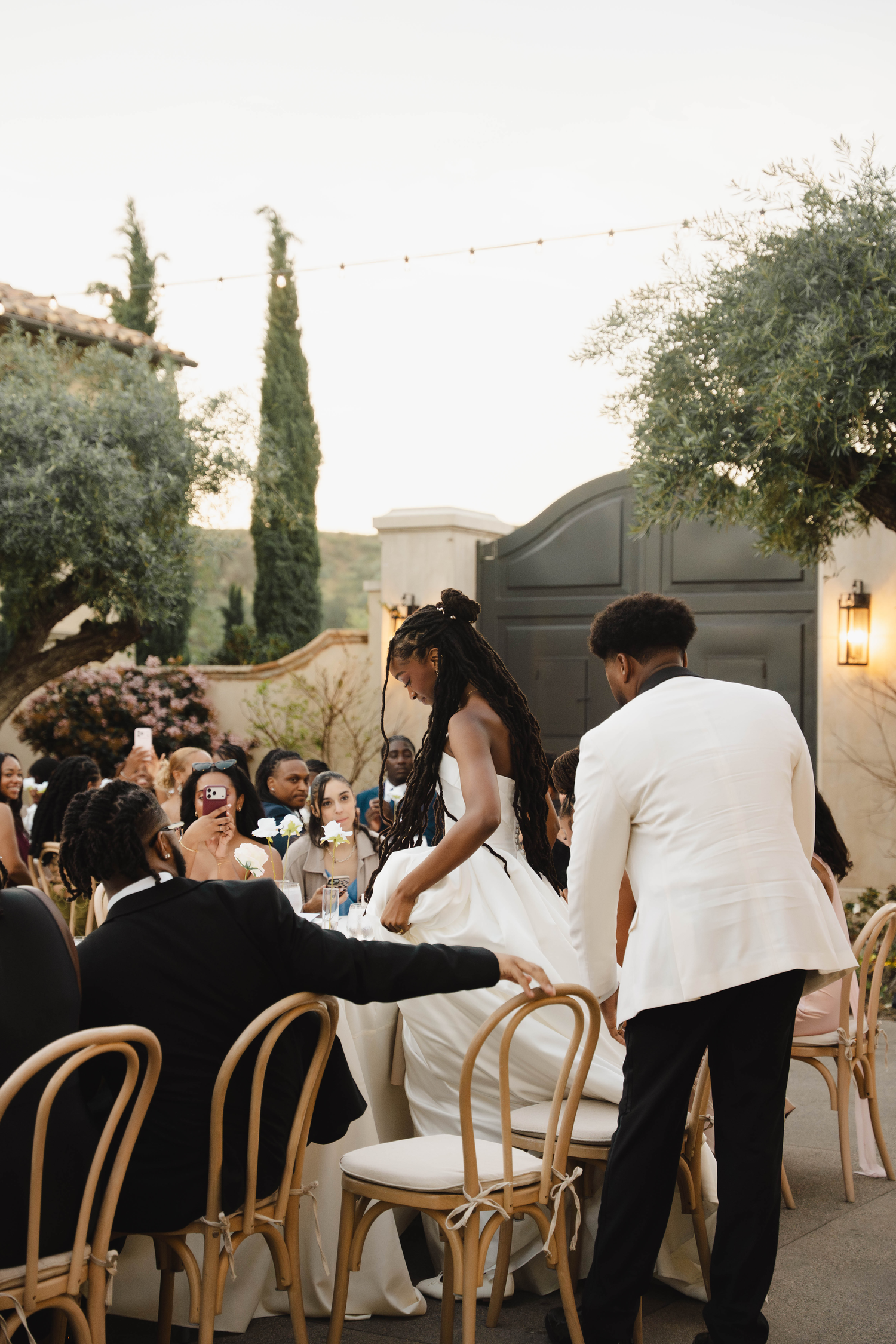 Bride in white gown assisted by groomsmen at outdoor wedding ceremony with guests seated in wooden chairs.