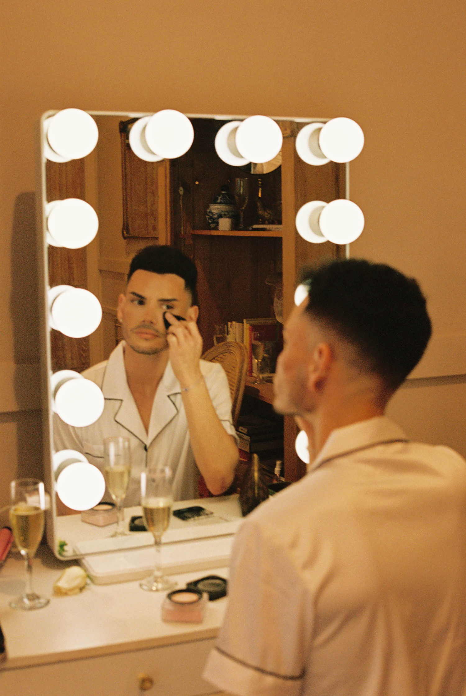 Man applying eye makeup at a Hollywood vanity mirror with bright bulbs, champagne glasses nearby.