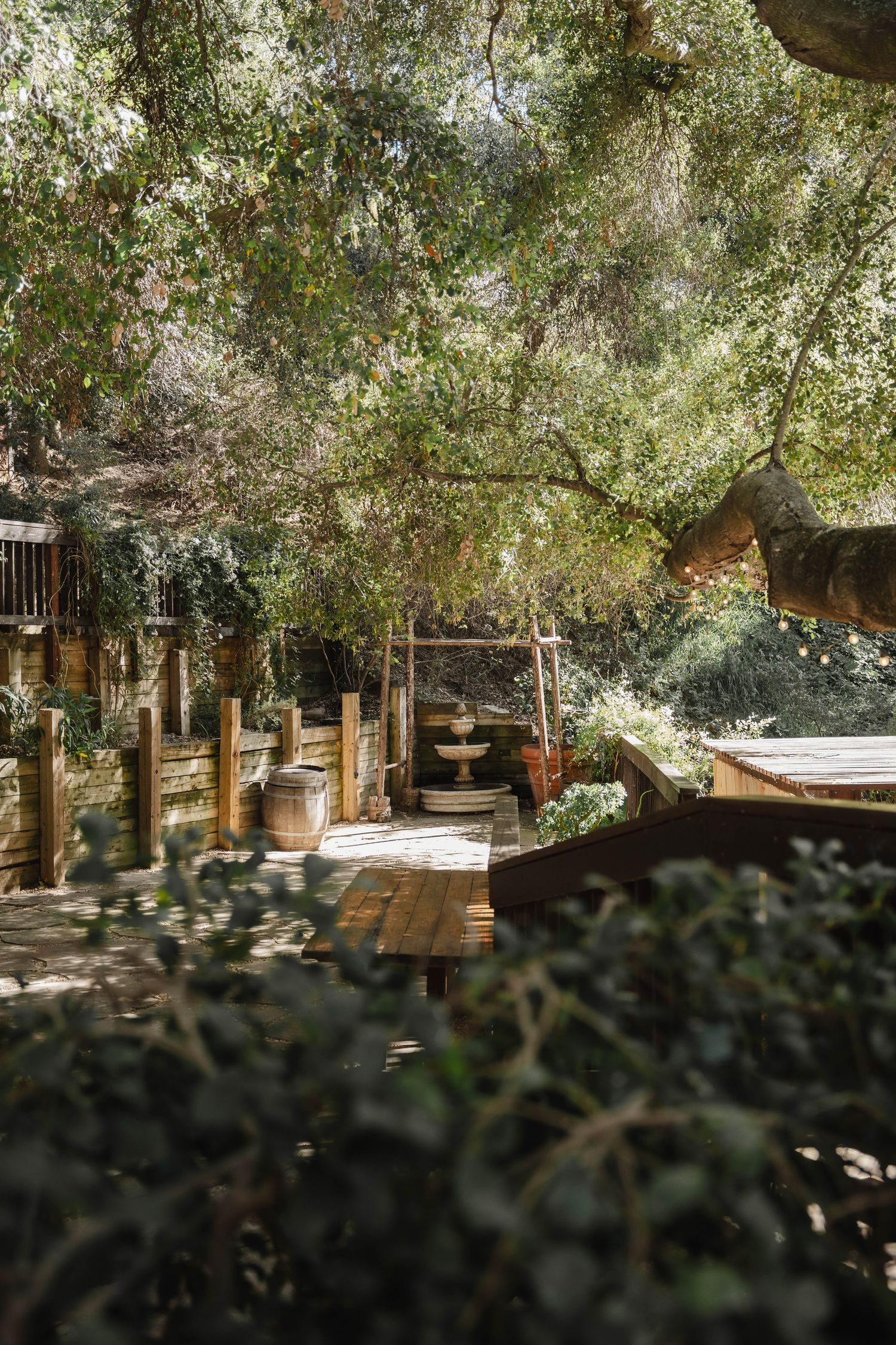 Rustic wooden deck surrounded by lush green trees with wine barrel planters and a stone fountain in a garden setting.