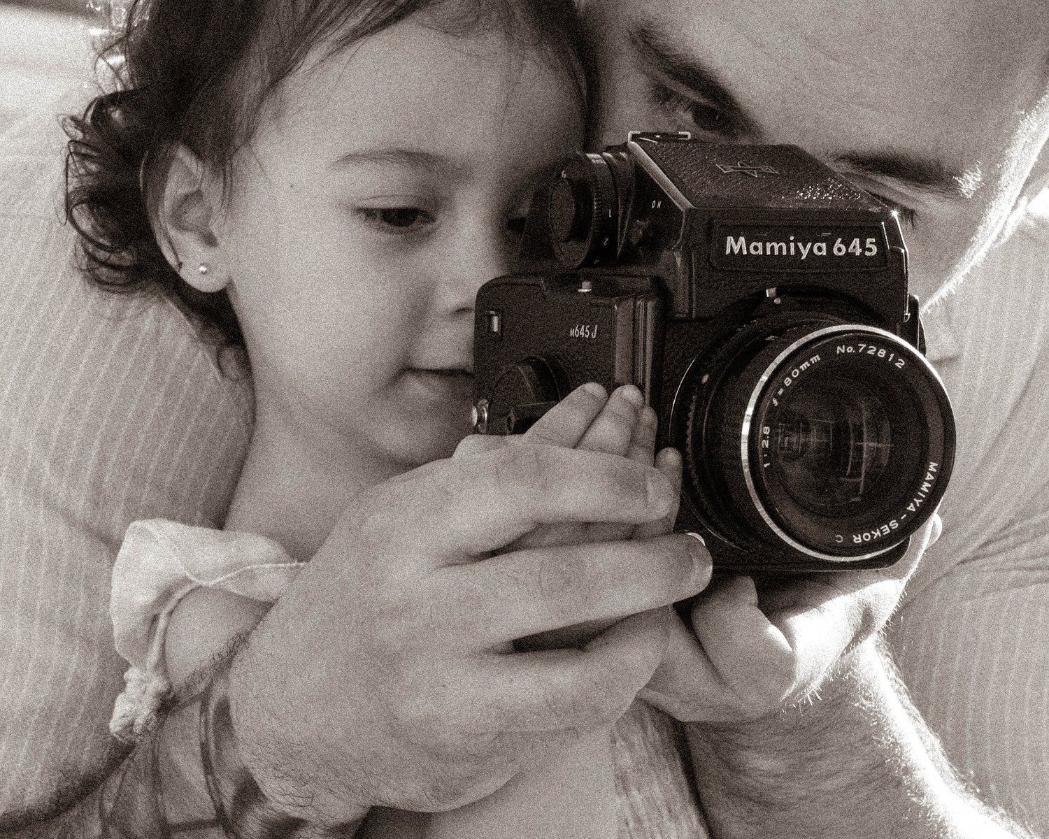 A toddler curiously peers into a Mamiya 645 medium format film camera held by an adult in a black and white photo.