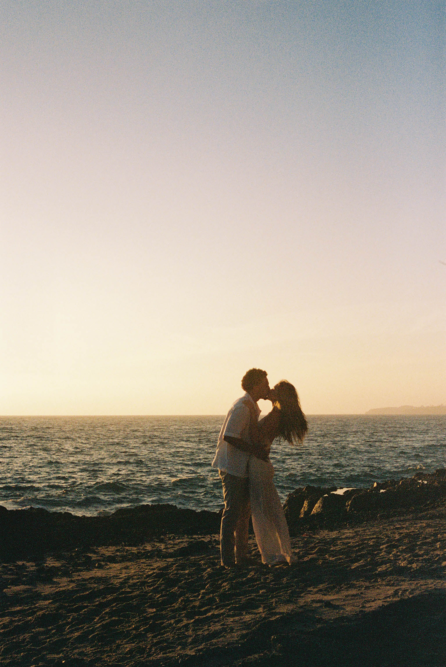 Romantic couple silhouetted kissing on rocky beach during golden sunset by the ocean.