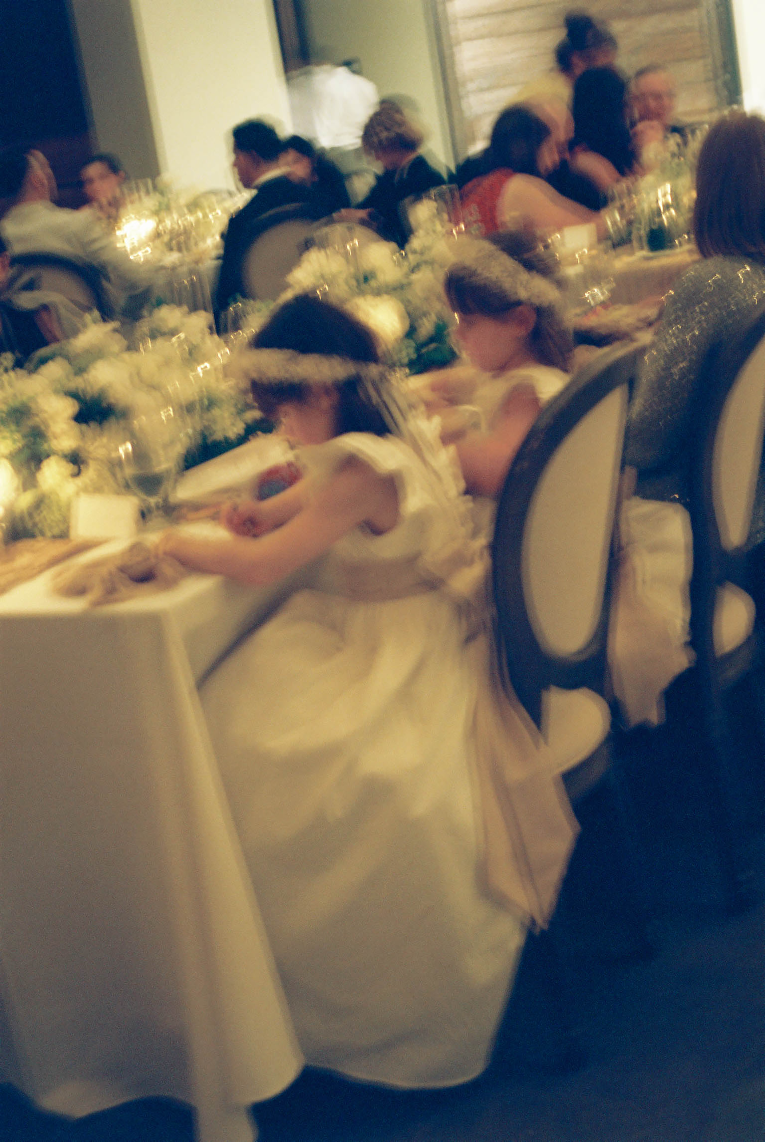 Blurred wedding reception scene with bride in white gown seated at elegant table with floral centerpieces and guests dining.