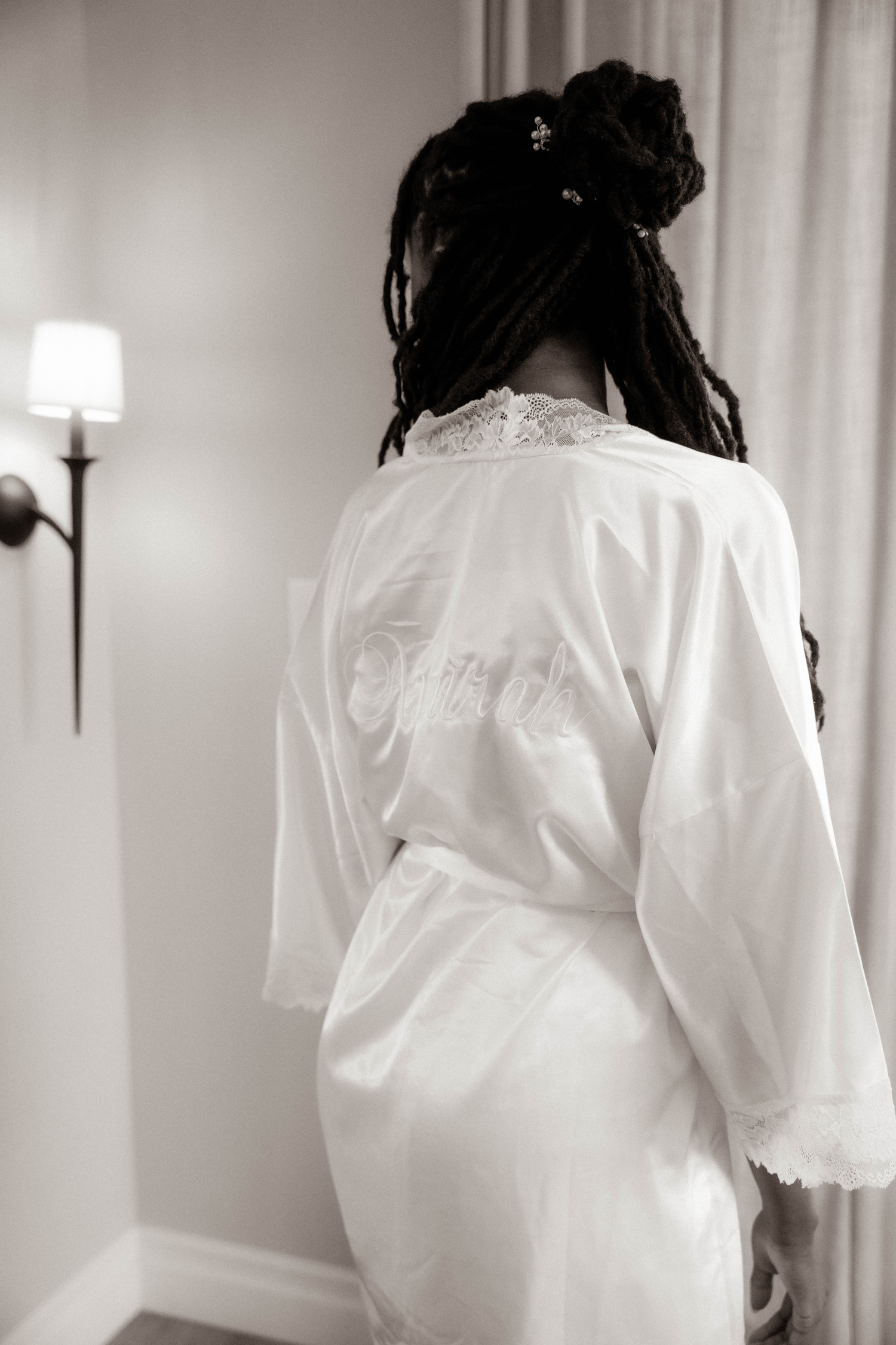 Black and white photo of a bride in a white lace robe with elegant updo hairstyle, seen from behind.