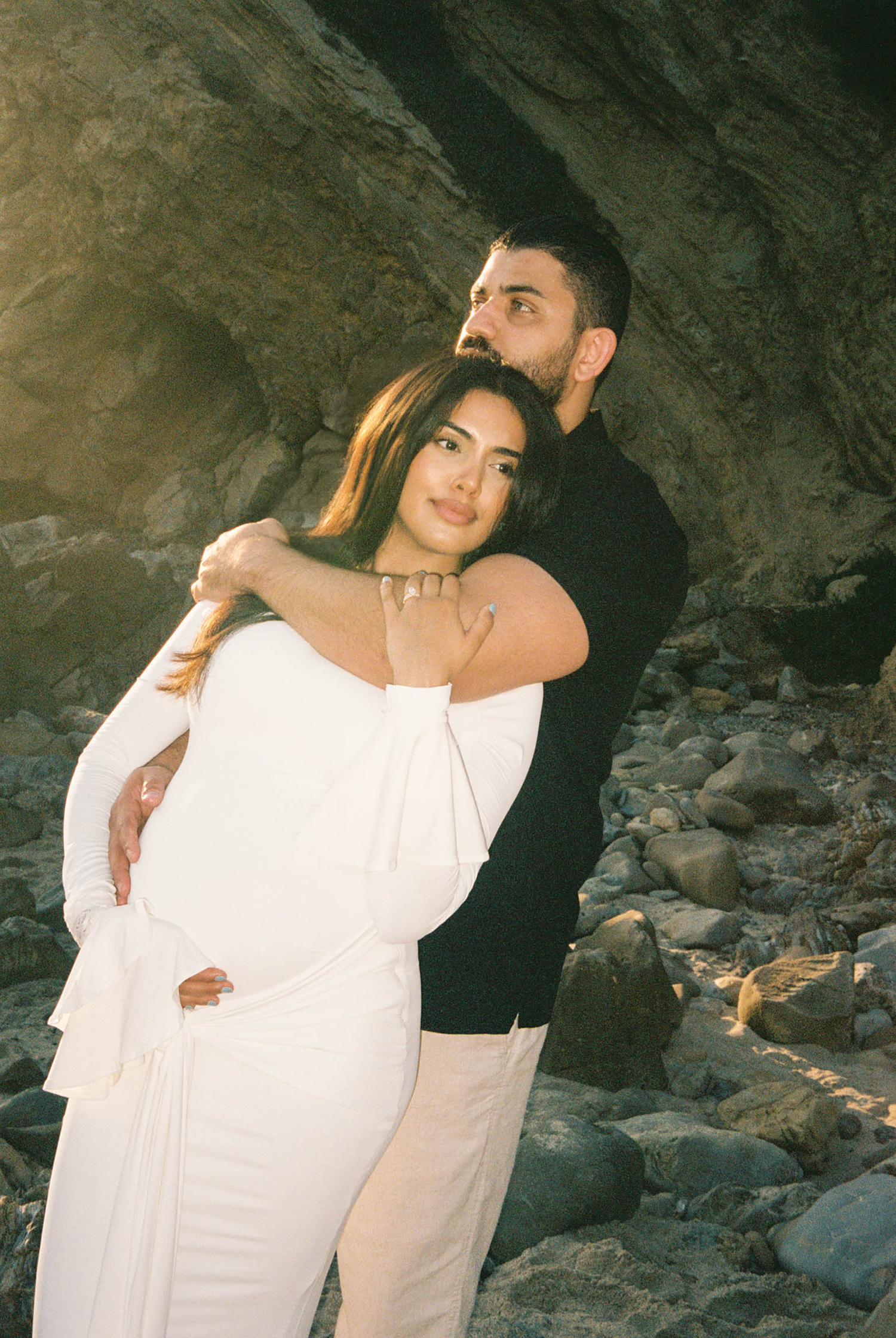 A couple embraces warmly against rocky cliffs at golden hour, woman in white off-shoulder gown.