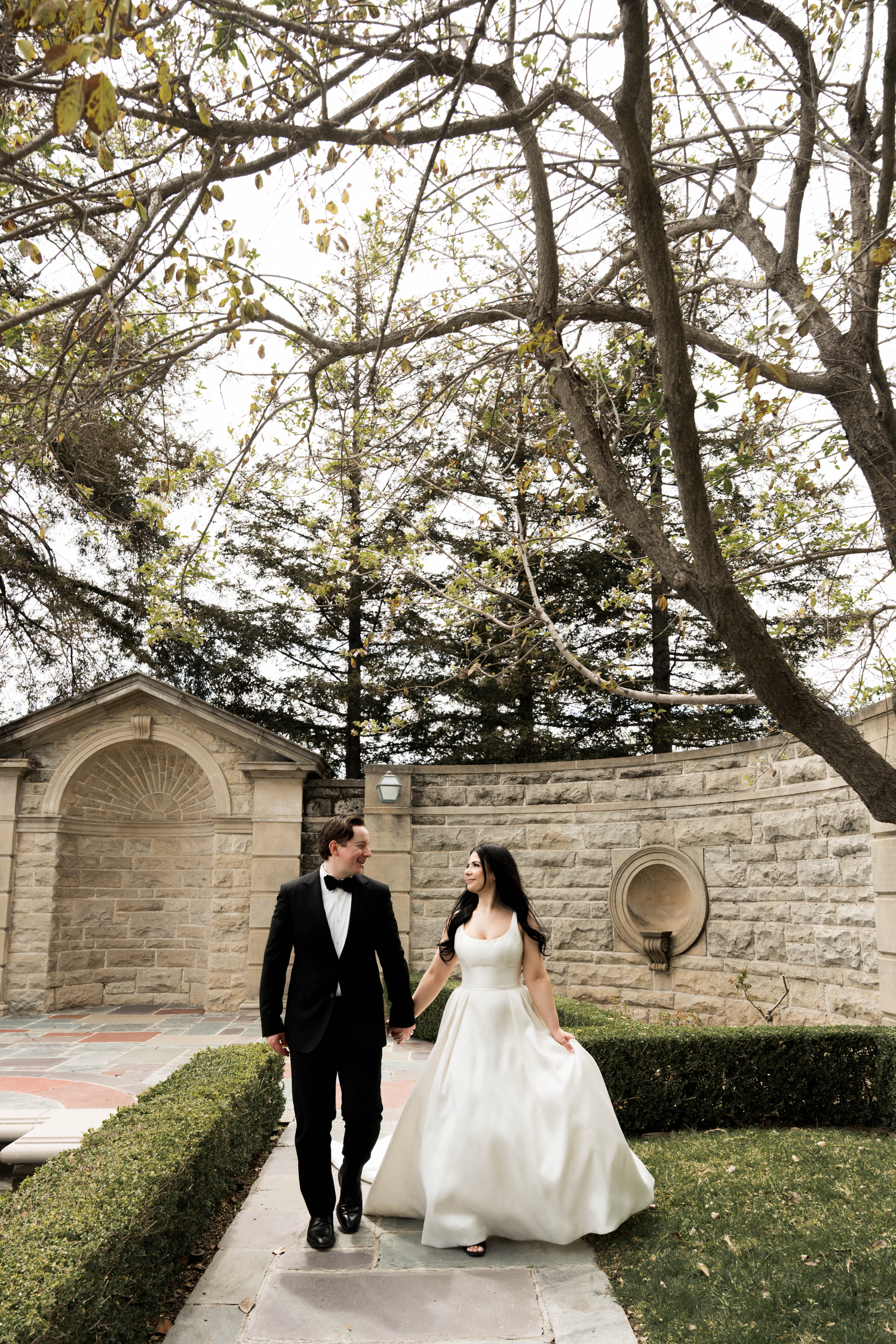 A bride and groom walk hand in hand through a garden pathway, she in a white gown, he in a black tuxedo.