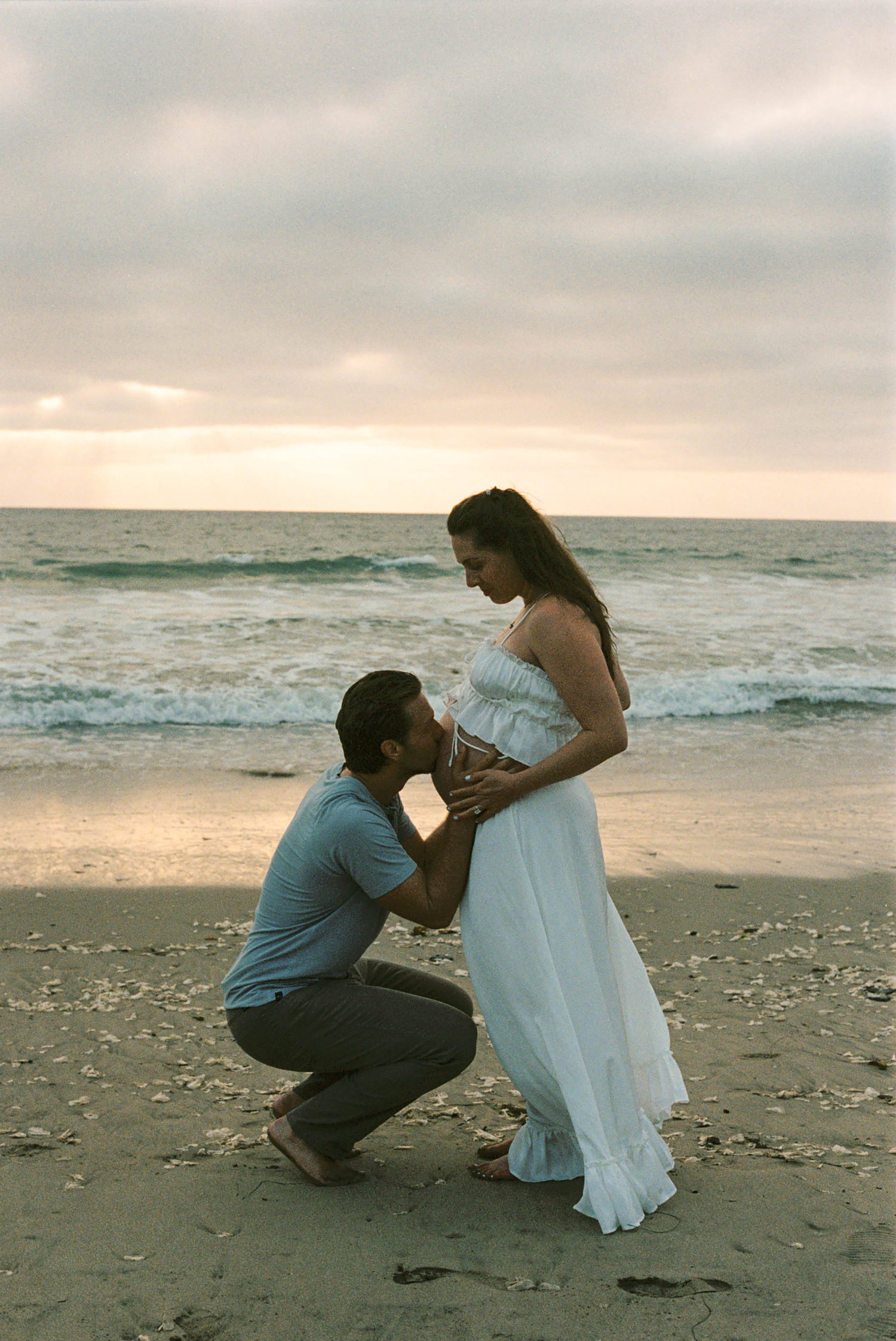 A man kneels on a beach at sunset, kissing a pregnant woman's belly as waves crash behind them.