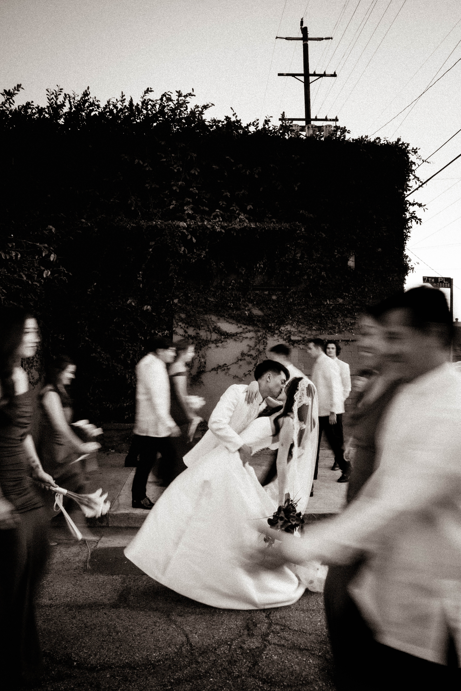 Black and white photo of bride and groom sharing a dip kiss surrounded by wedding party outdoors at dusk.