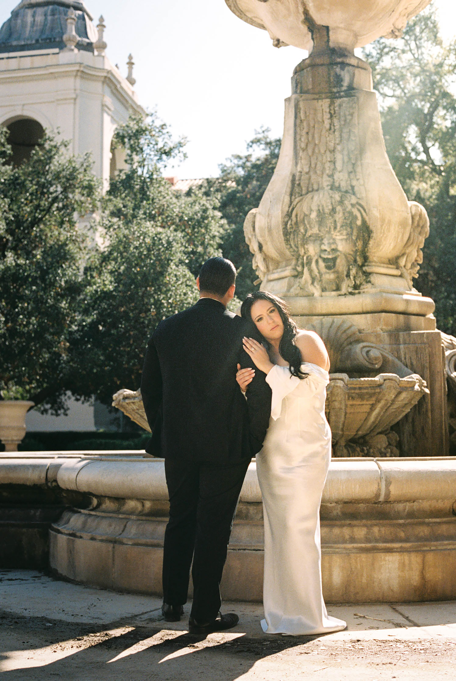 Bride in white gown embraces groom in black suit by ornate stone fountain in sunlit garden.