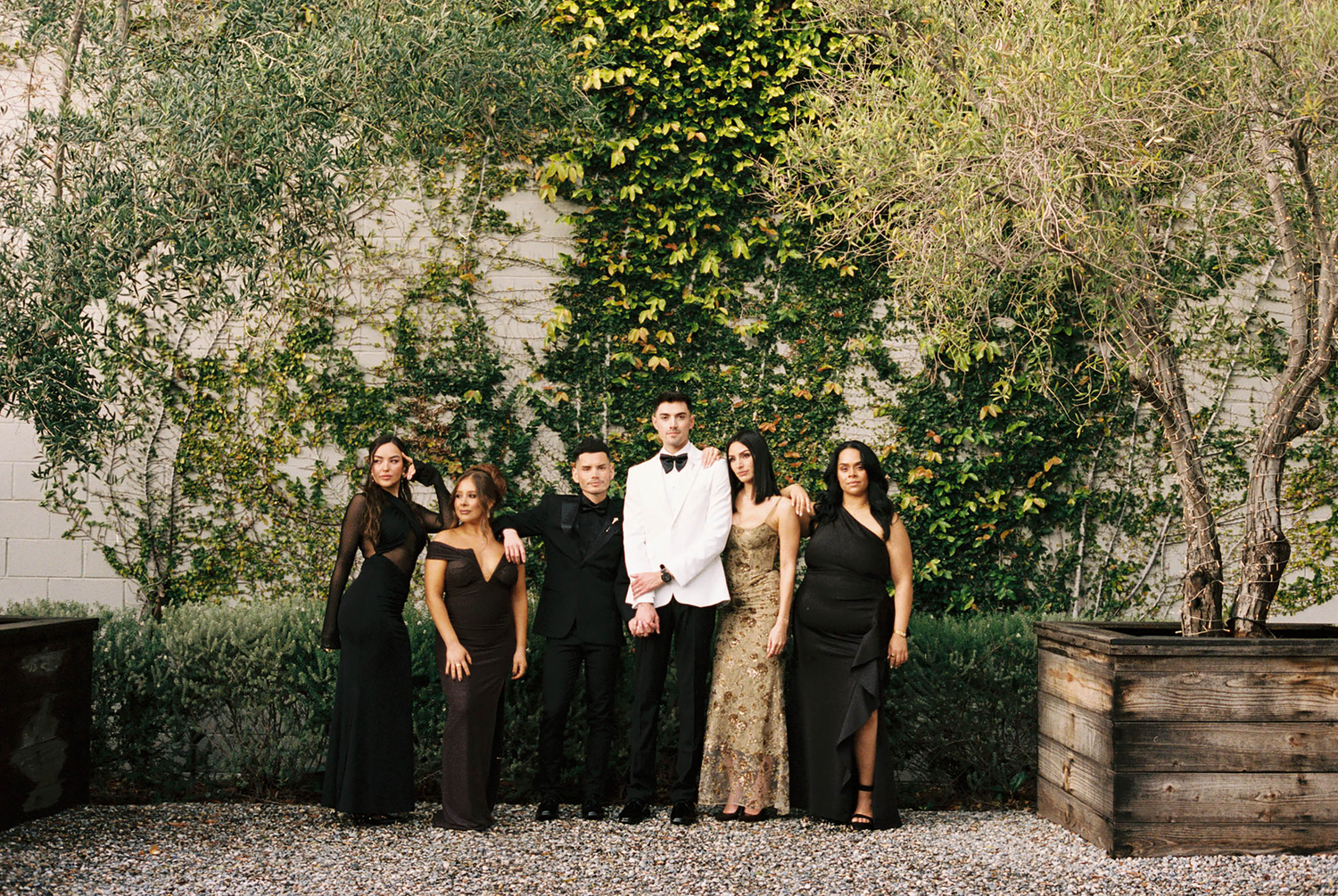 Group of elegantly dressed people posing outdoors in black formal wear with one person in white tuxedo.