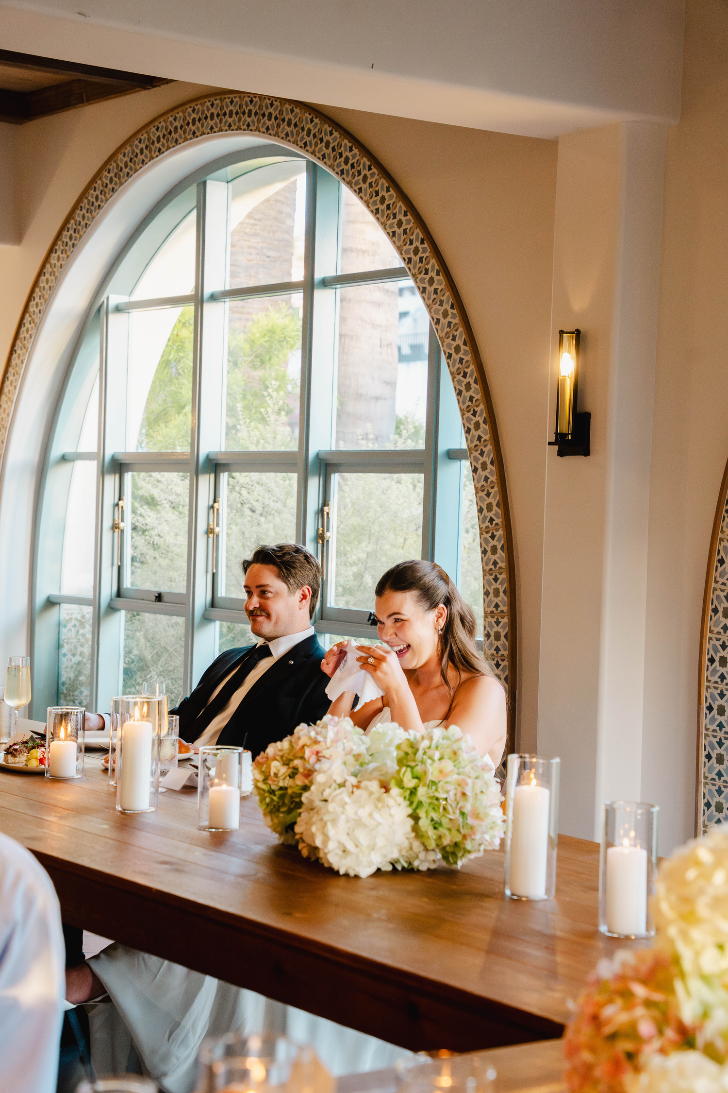 Bride and groom seated at wedding reception table with white floral centerpieces and candles beneath an arched window.