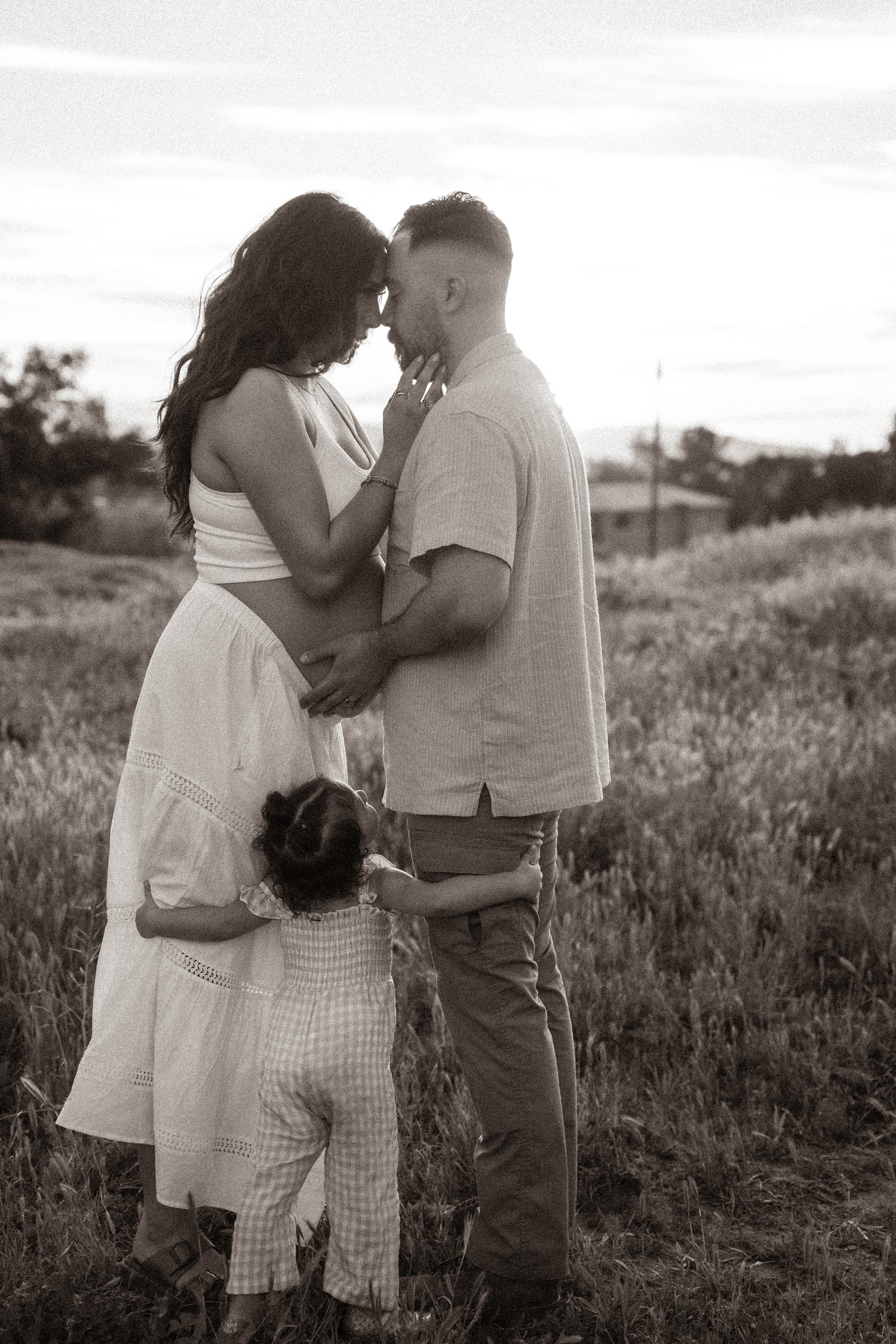 Black and white photo of a couple sharing a tender moment in a field while their young child embraces them.