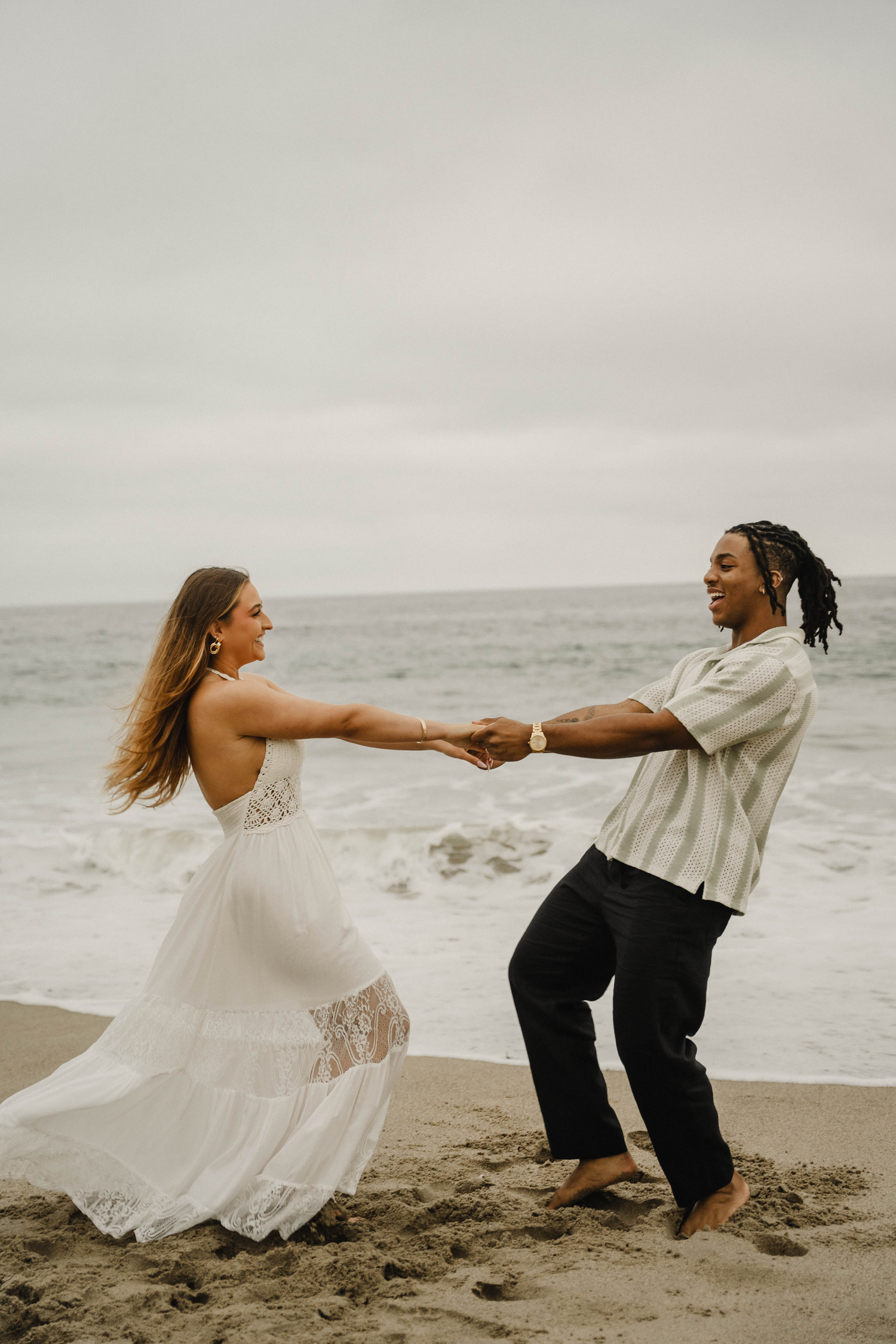 A joyful couple dances on a sandy beach, woman in white lace dress, man in striped shirt, ocean waves behind them.
