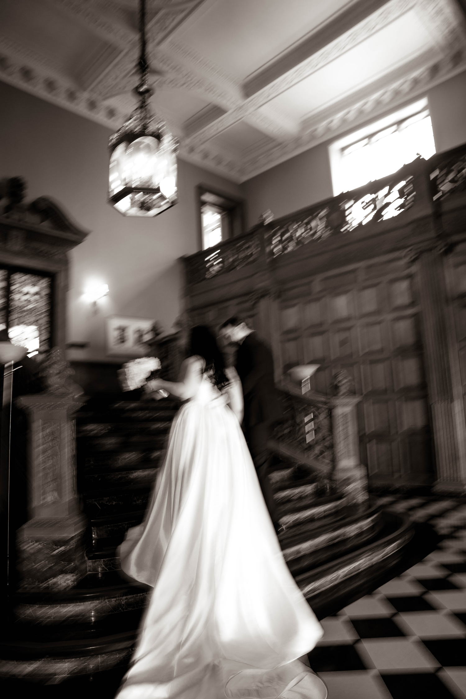 Black and white photo of bride and groom in elegant mansion foyer with grand staircase and chandelier.