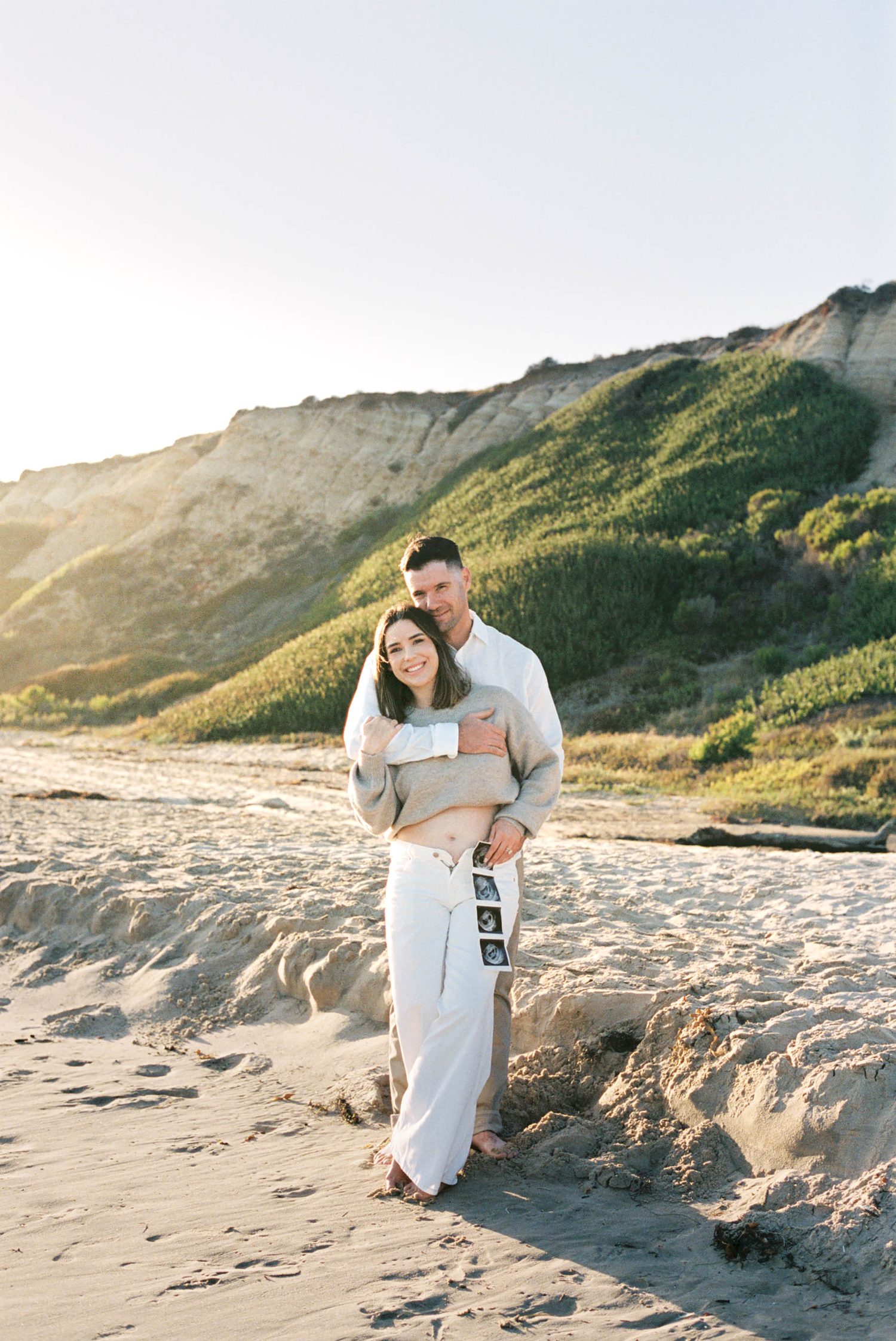 Couple embracing on a beach at sunset, holding ultrasound photos, with green cliffs in the background.