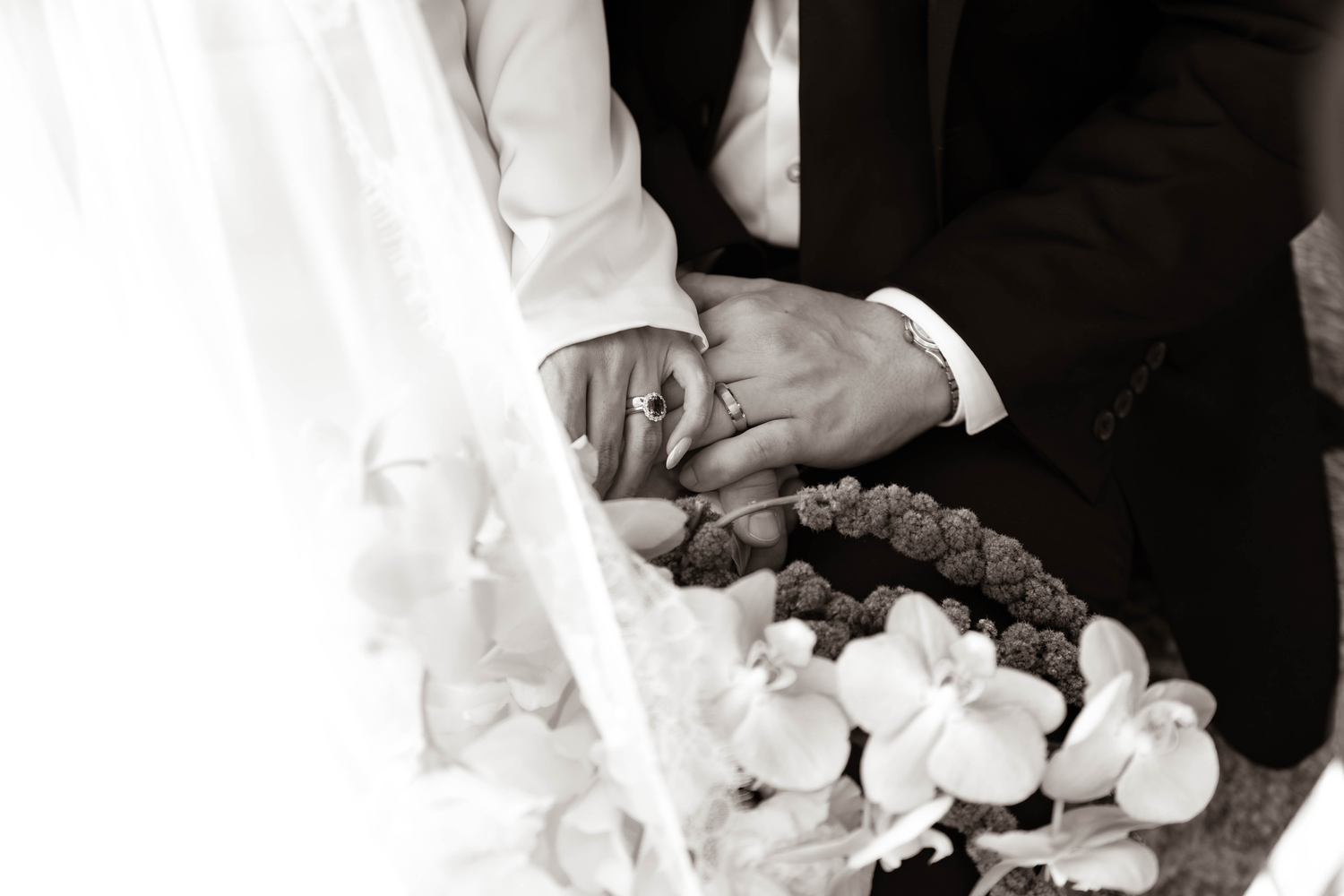 Black and white wedding photo of bride and groom holding hands with floral arrangement and wedding rings visible.