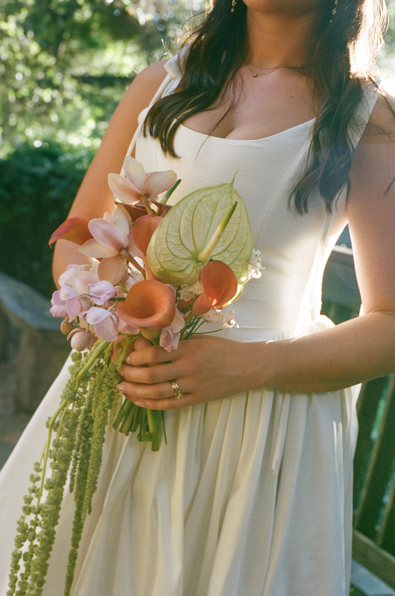 Bride in white dress holding tropical wedding bouquet with anthurium, pink orchids, and coral calla lilies outdoors.