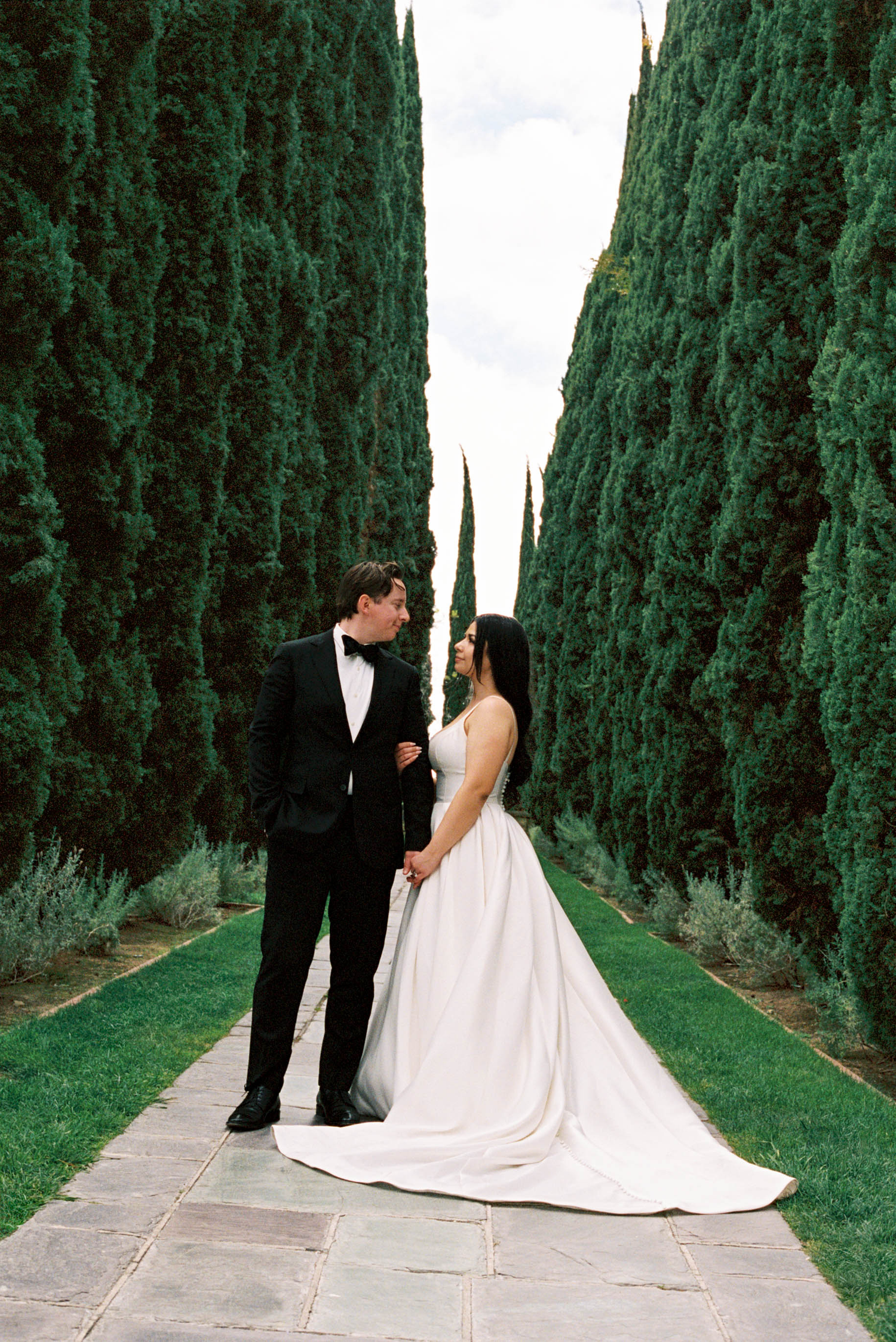 Bride and groom stand on stone path between tall cypress trees, bride in white gown, groom in black tuxedo.