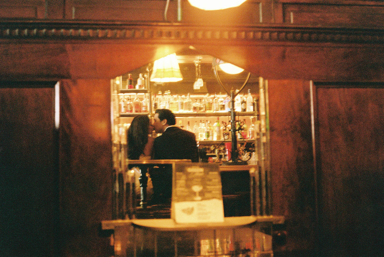Couple reflected in bar mirror surrounded by warm ambient lighting and shelves of bottles.