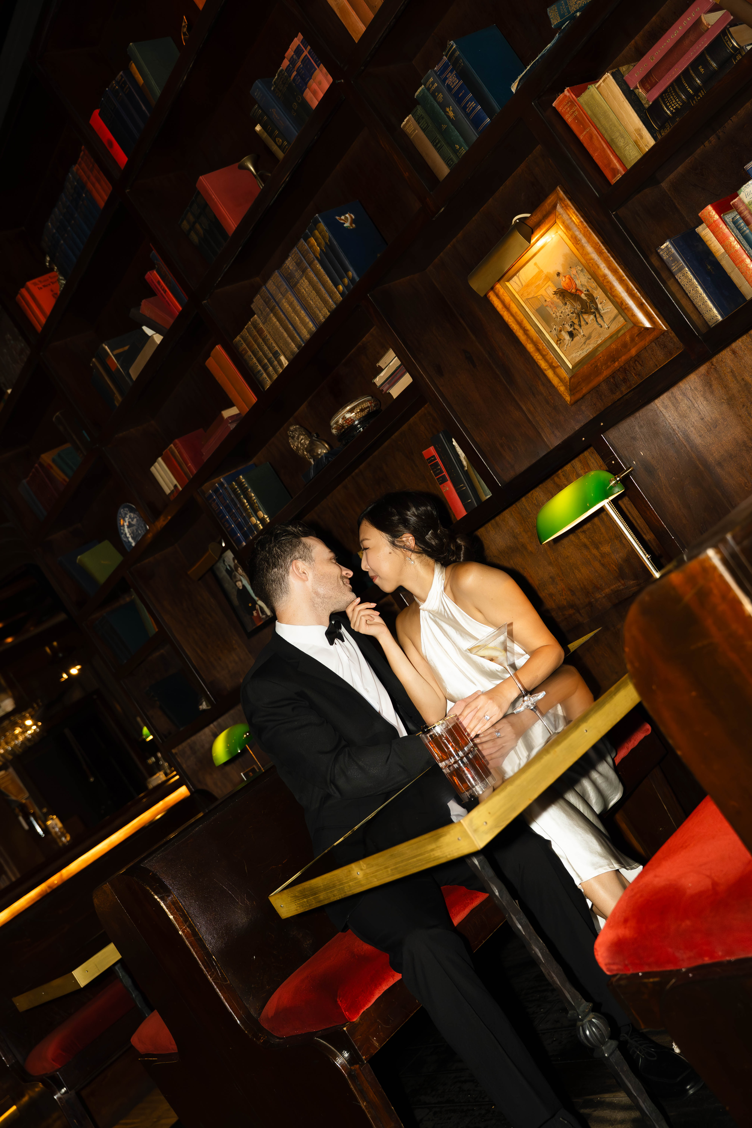 A couple in formal attire shares a romantic moment at a table in an elegant library bar with bookshelves.