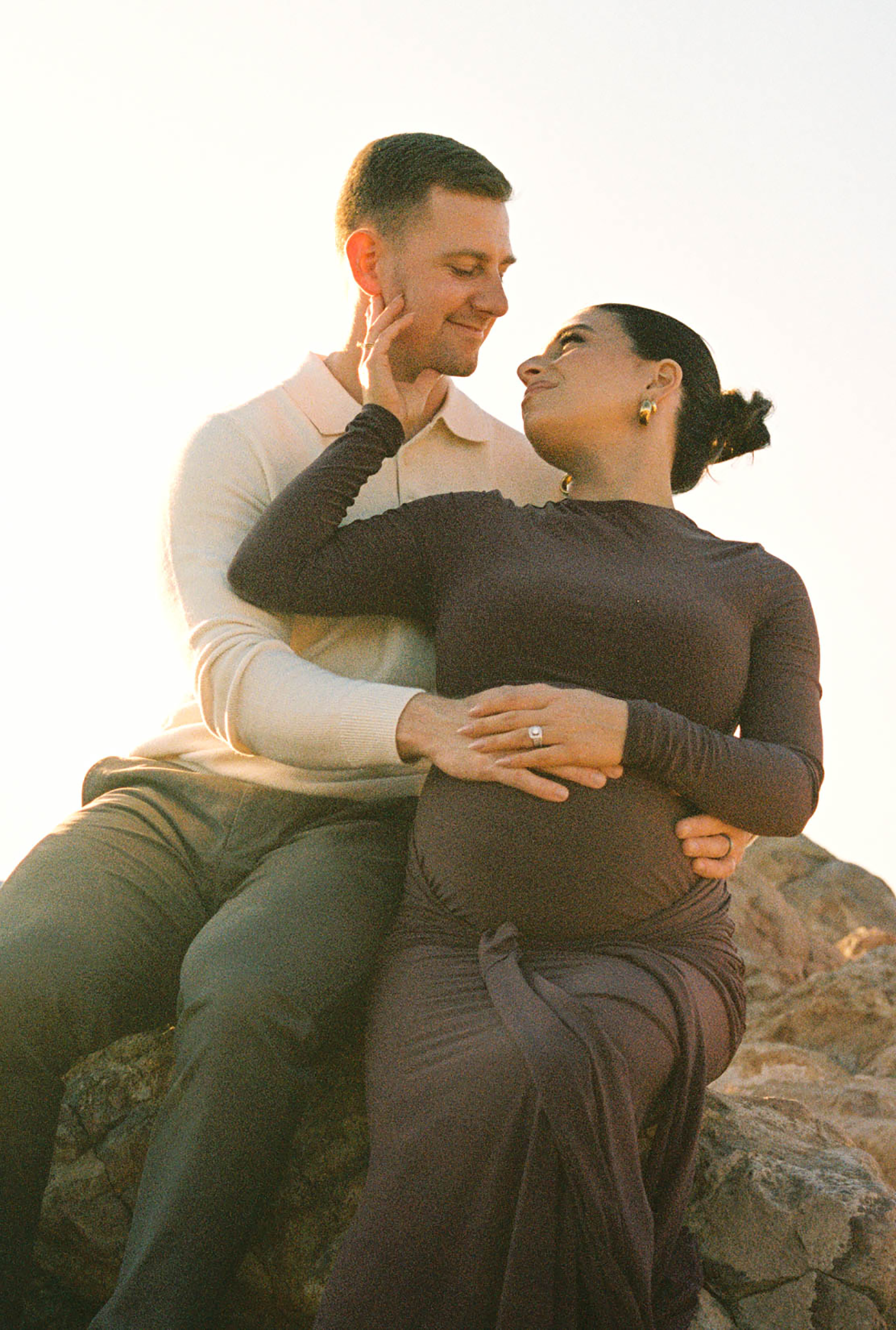 A couple shares a tender moment on rocks at sunset, with the woman visibly pregnant, wearing a brown dress.