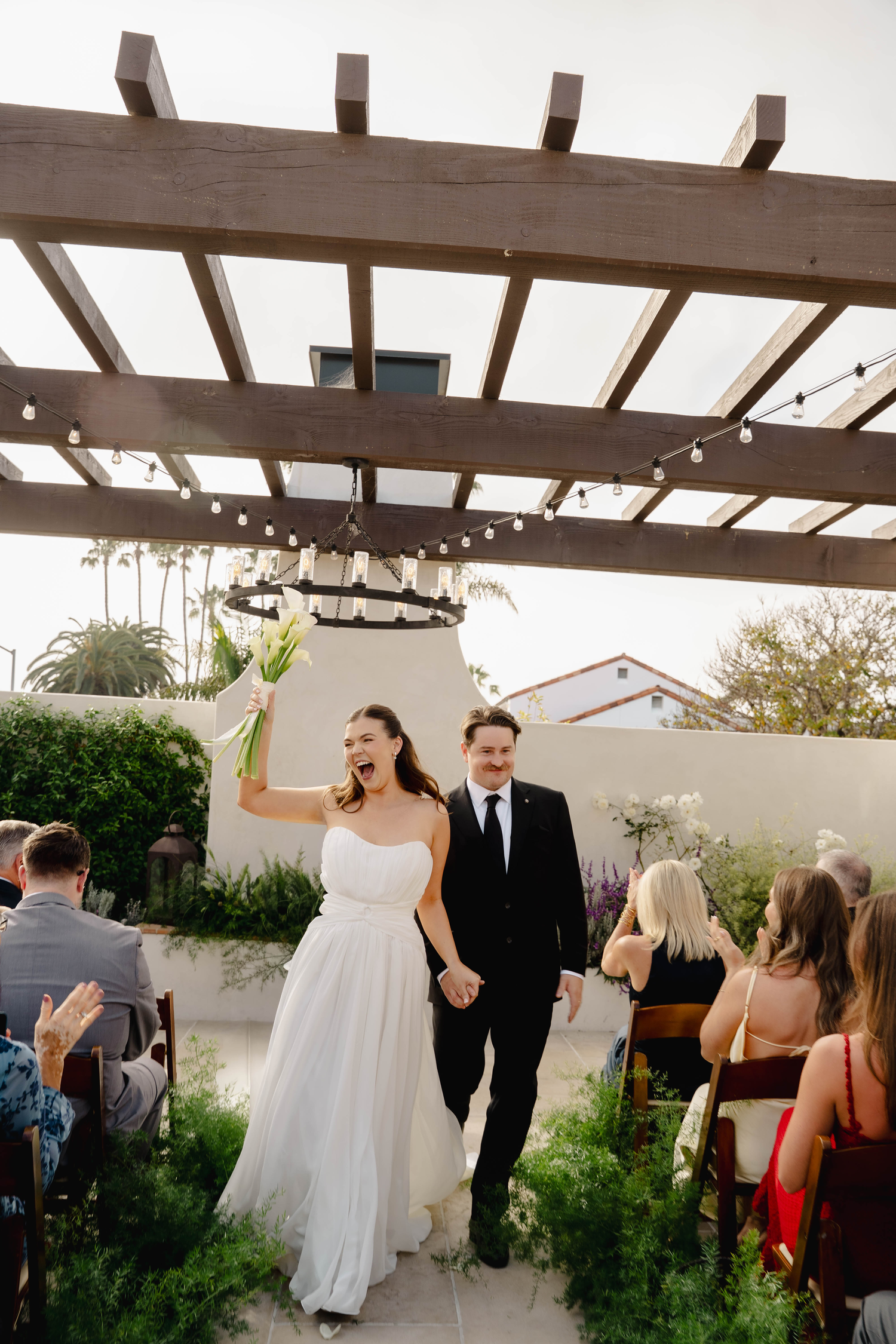 Joyful bride and groom walk down outdoor aisle after wedding ceremony under wooden pergola with string lights.