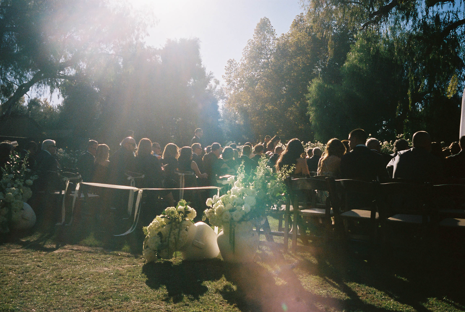 Outdoor wedding ceremony in a sunlit garden with guests seated on wooden benches surrounded by lush green trees.