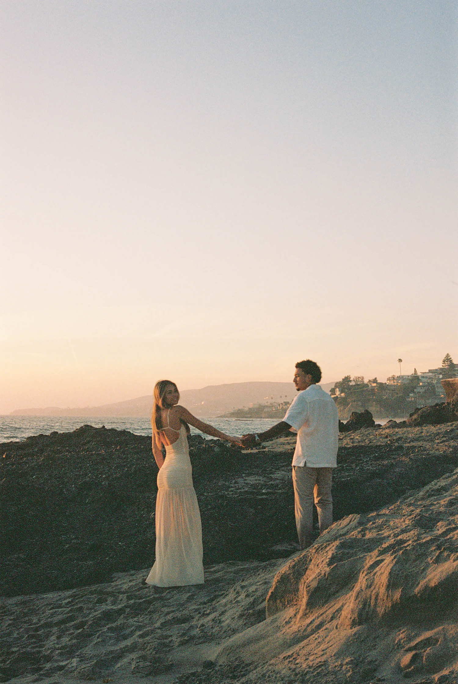 Couple holding hands on rocky coastal cliffs during a warm pastel sunset by the sea.